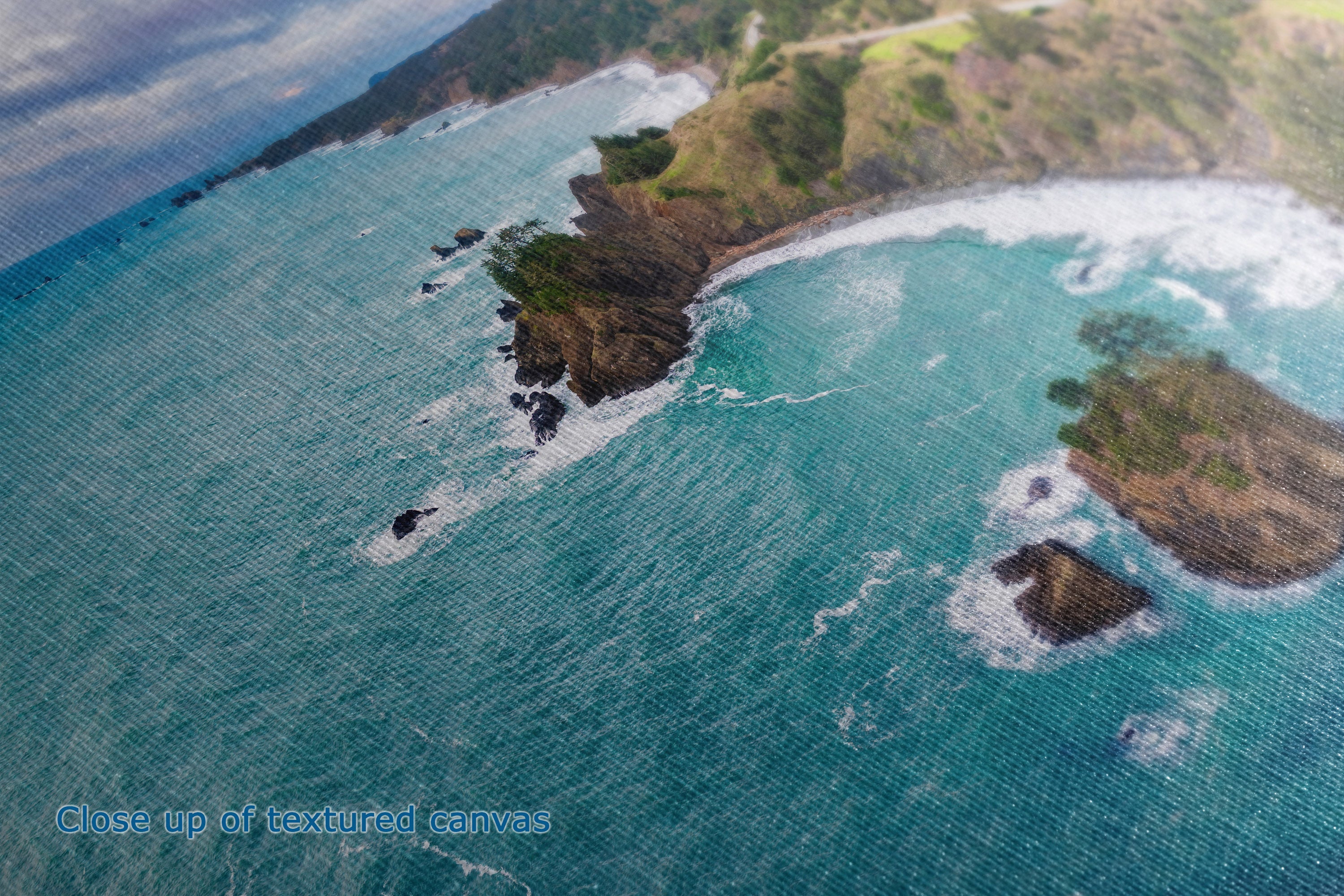 A close up view of a canvas wall art print showing the detail and texture of an aerial view of a rocky coastline with turquoise ocean water, white surf, and green cliffs. A small blue text watermark at the lower left reads 'Close up of textured canvas.