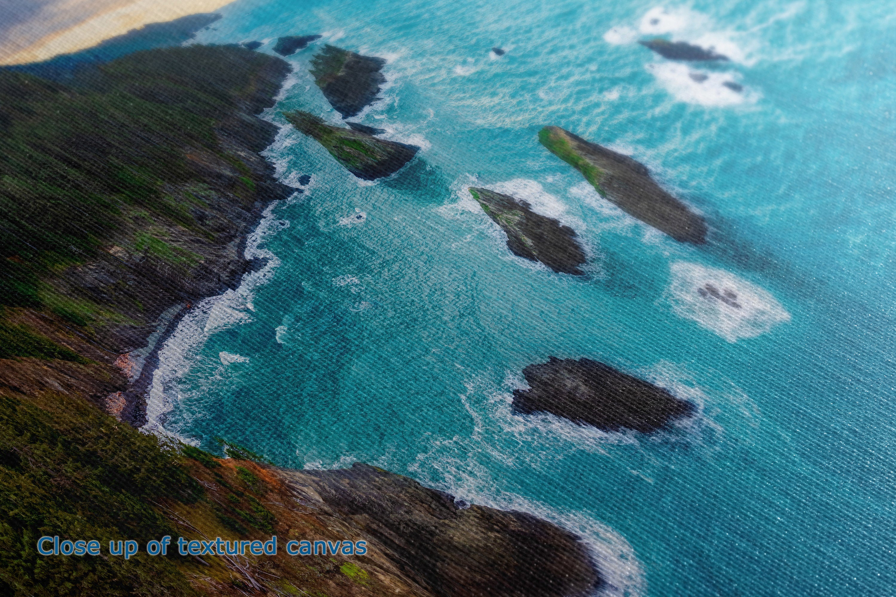 A close-up of a textured canvas print depicting an aerial coastal scene with turquoise ocean water, white surf, dark rocky islands, and green-brown cliffs. Blue text in the lower-left corner reads 'Close up of textured canvas'.
