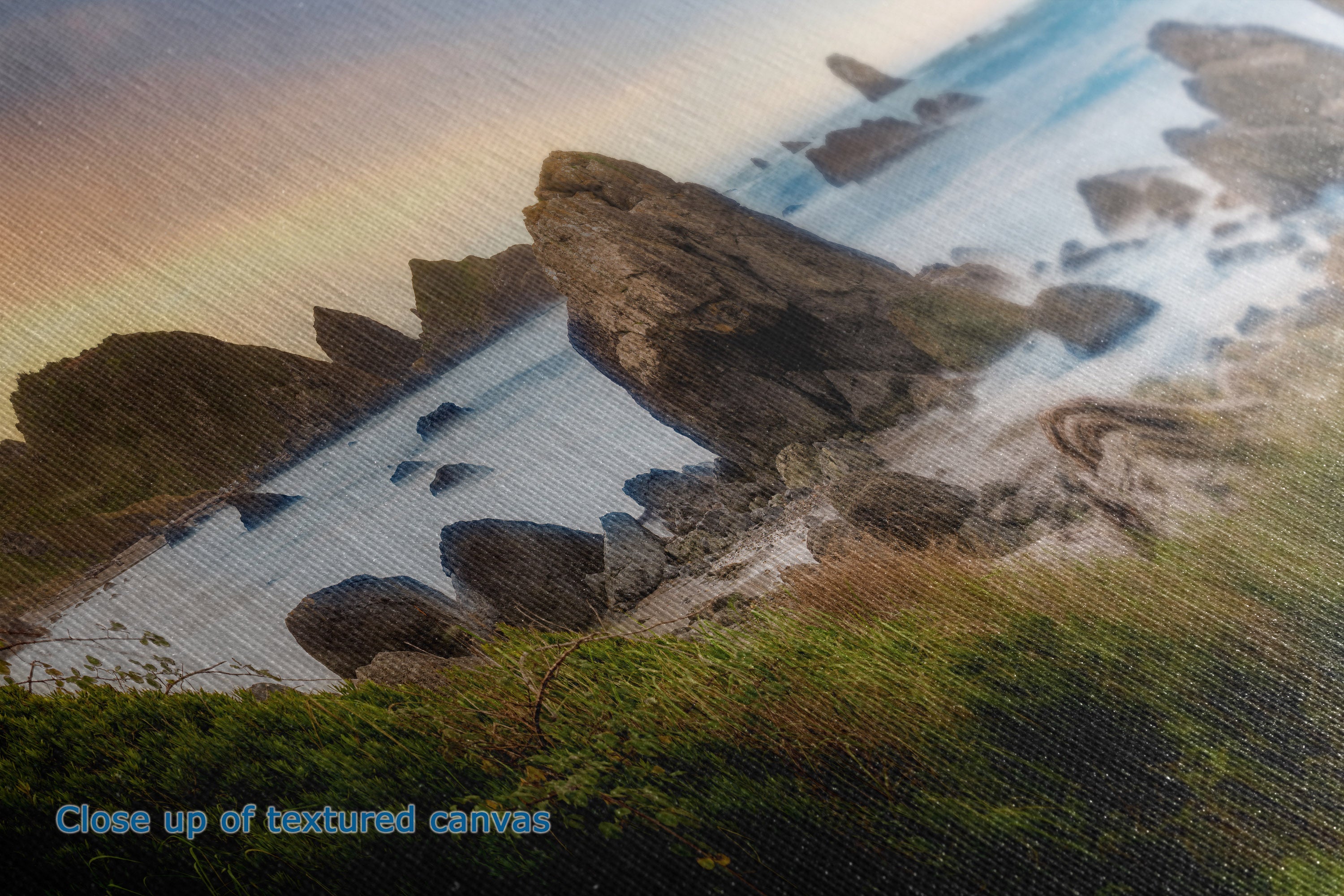 Close-up of a textured canvas print depicting a rocky coastline with a bay, green grass in the foreground, and a warm sunset sky. Blue text in the lower-left corner reads 'Close up of textured canvas'.