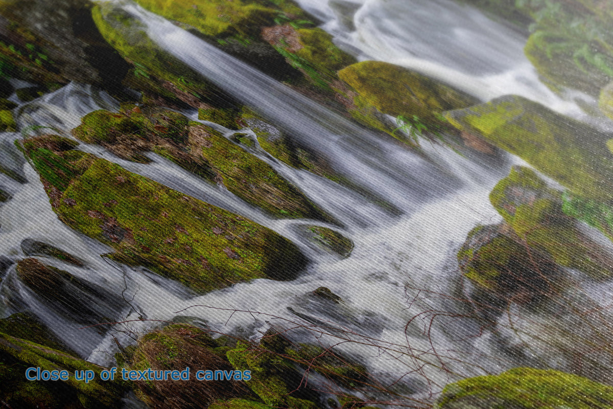 Close-up of a textured canvas print depicting a scenic waterfall flowing over mossy green rocks, with a blue text overlay in the bottom-left corner reading 'Close up of textured canvas'.