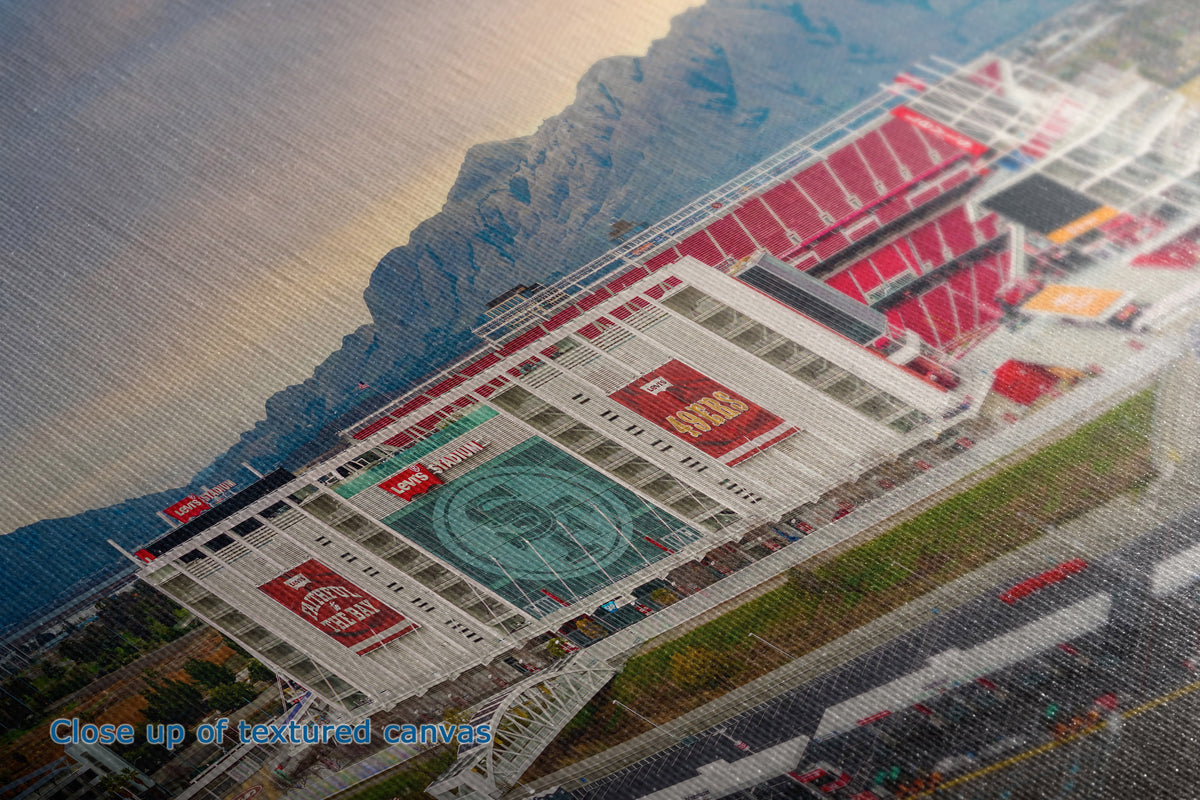 Close-up of the detail and texture of a canvas print depicting an aerial view of a stadium with red seating, green fields, and mountains in the background, with a blue text overlay reading 'Close up of textured canvas'.