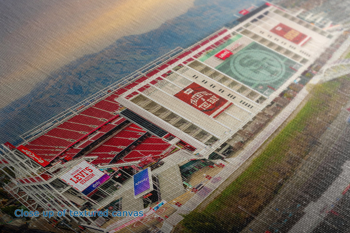 A close-up of the detail and texture of a  canvas print depicting an aerial view of a stadium with red seating, green fields, and surrounding hills under a sunset sky. A blue text overlay at the bottom left reads 'Close up of textured canvas'.