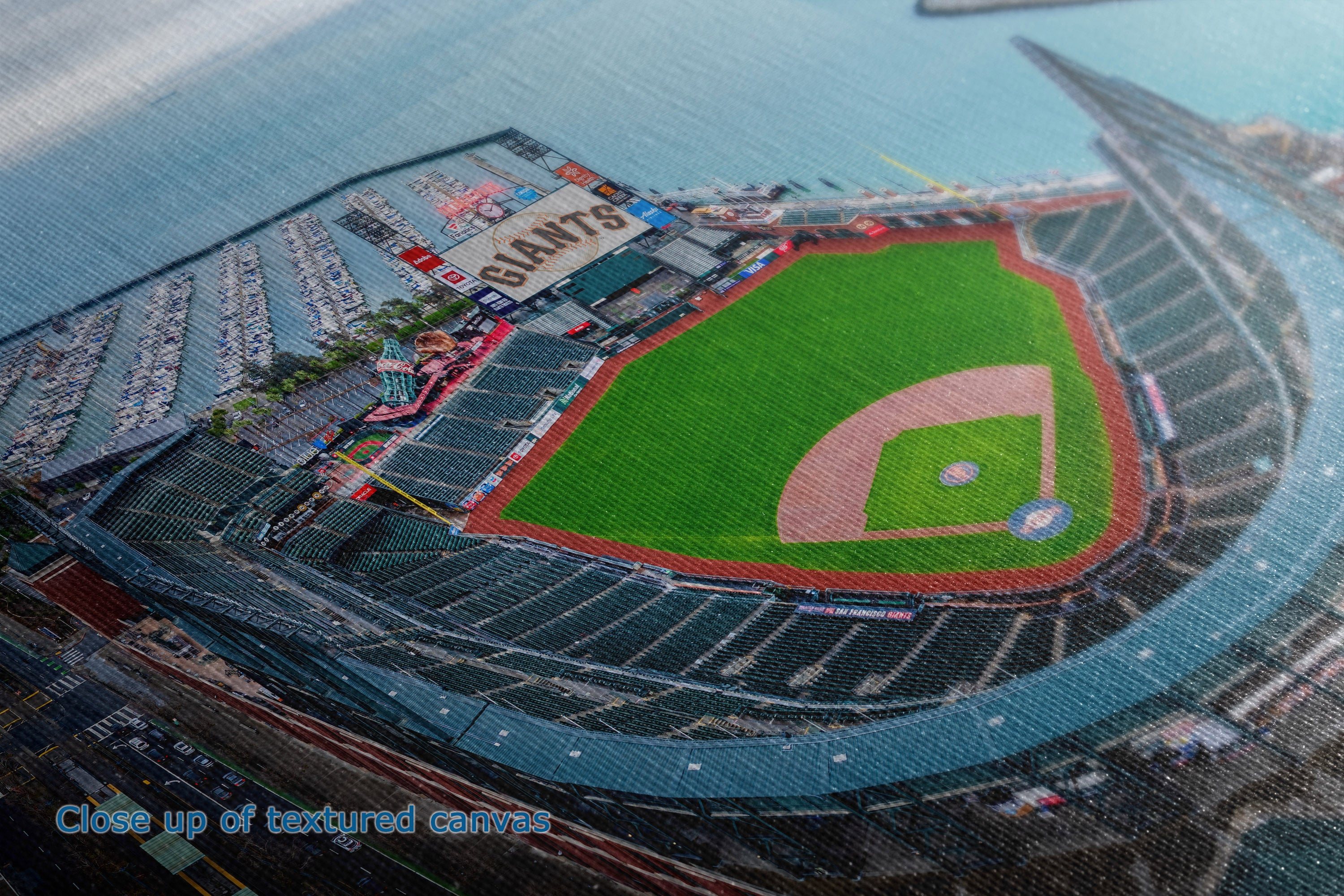 A close-up detail and texture view of a canvas wall art print showing a baseball stadium interior and field, with the word 'GIANTS' visible on the scoreboard. The photo shows the green infield, dirt pitching area, seating tiers, and surrounding blue-gray stadium structure.