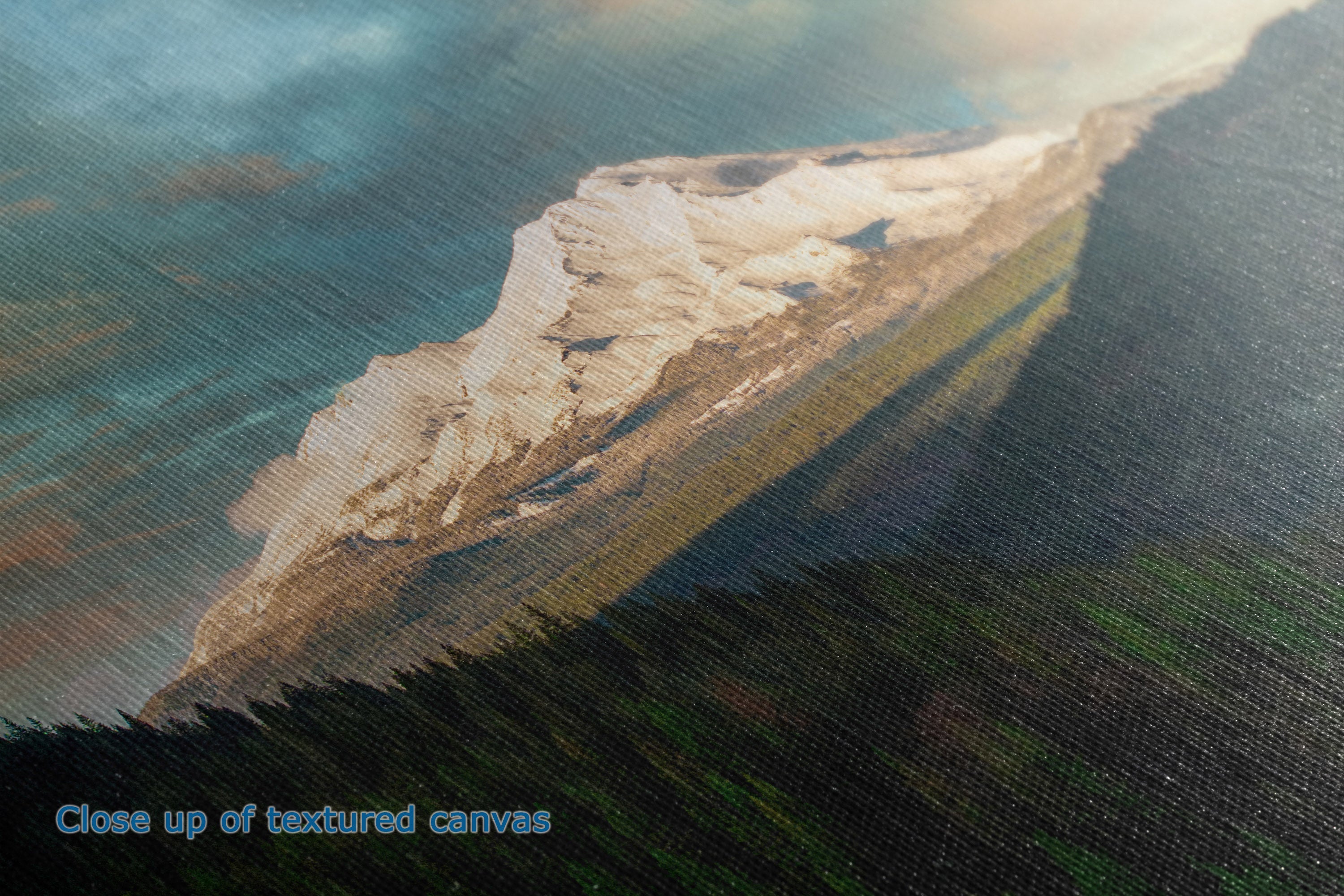 Close-up of a textured canvas photographic wall art depicting a snowy mountain peak rising above dark green forested hills under a cloudy sky.