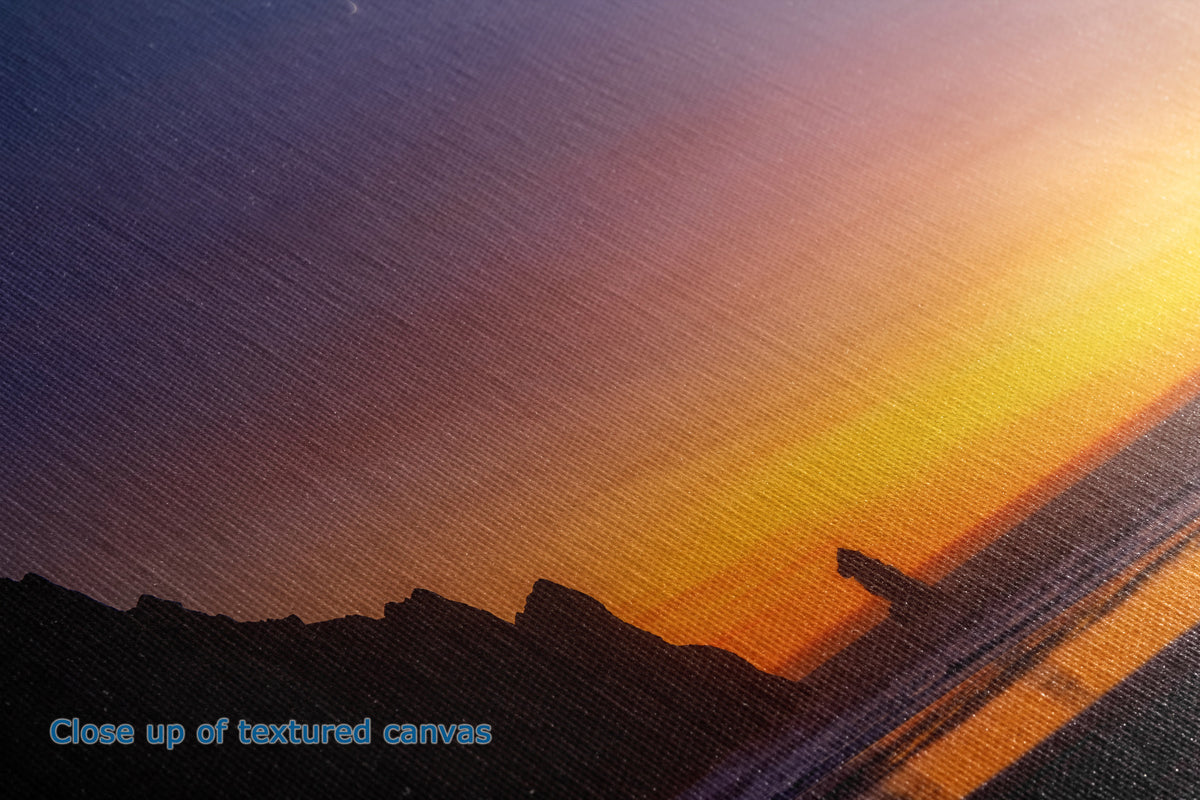 Close-up of a textured canvas print depicting a sunset over a shoreline silhouette, with a blue text overlay reading 'Close up of textured canvas' in the lower-left corner.