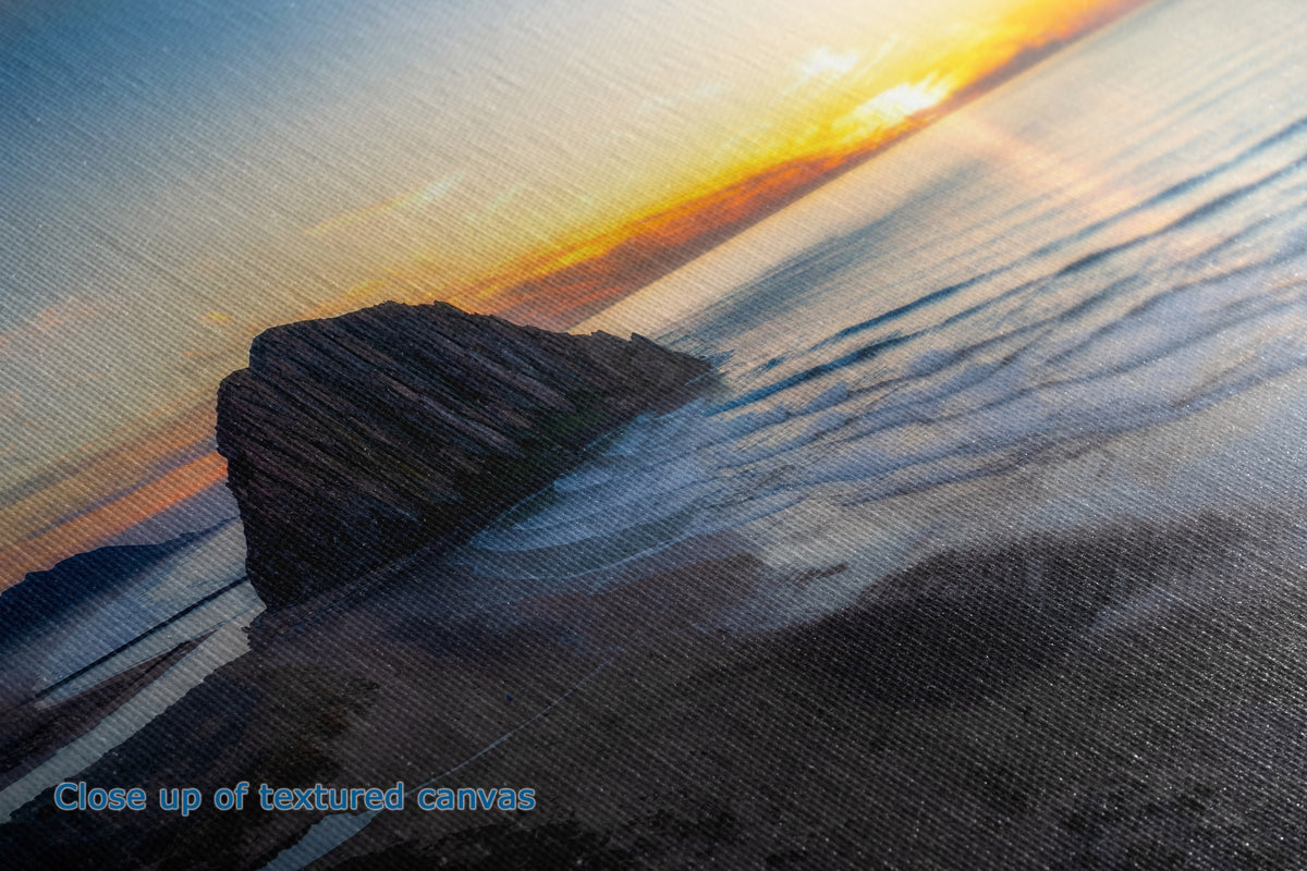 Close-up of a textured canvas print depicting a coastal sunset scene with a rock formation, ocean waves, and warm golden light on the horizon.