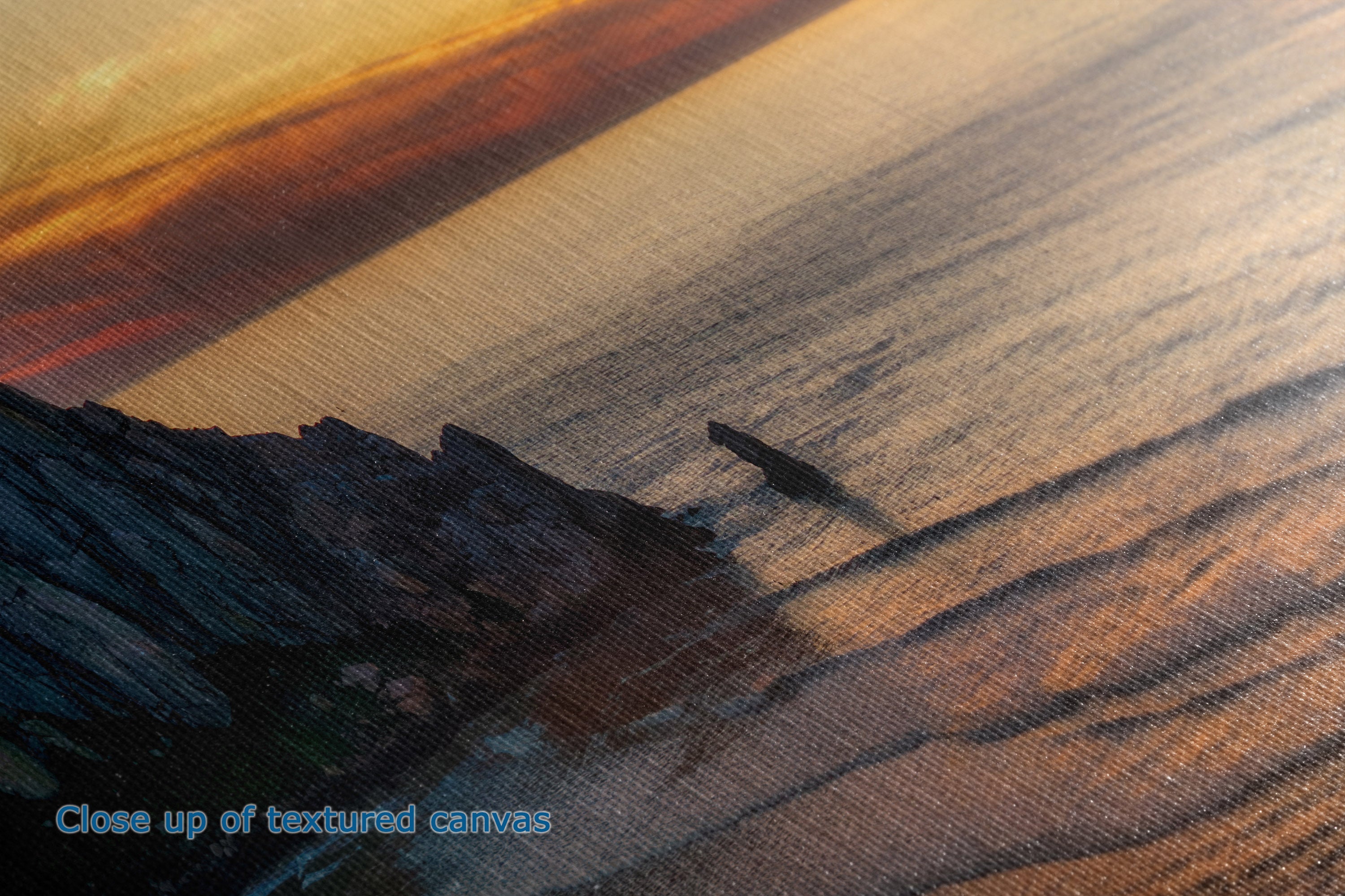 Close-up of a textured canvas print depicting a dramatic sunset over a rocky coastline with a small boat on the water, showing warm orange and golden light reflections.