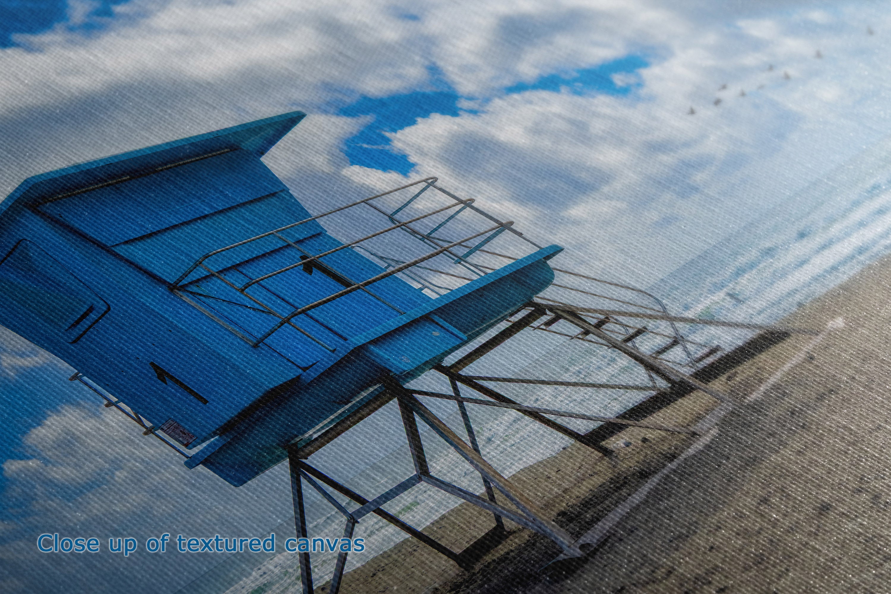 A close-up of a textured canvas print depicting a blue lifeguard tower on a sandy beach under a cloudy blue sky, with the text 'Close up of textured canvas' in the lower-left corner.