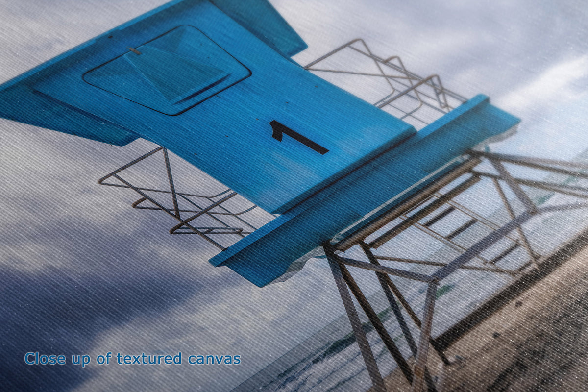 Close-up of a textured canvas print depicting a bright blue lifeguard tower against a cloudy sky and beach background, with the text 'Close up of textured canvas' in the bottom-left corner.