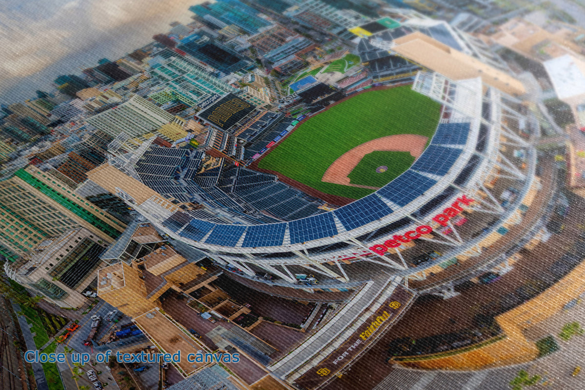 A close up view of the detail and texture of a canvas wall art print that depicts and aerial view of a baseball stadium with a green field, red dirt infield, and a curved roof covered in dark blue solar panels, surrounded by urban buildings. Large red text on the stadium reads 'PETCO PARK'.