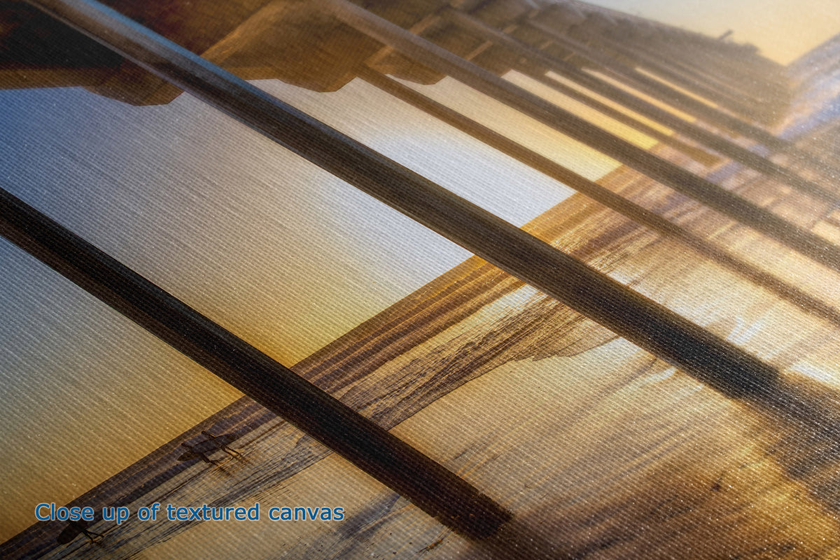 Close-up of a textured canvas print featuring a warm-toned sunset scene with diagonal wooden railings and golden light.