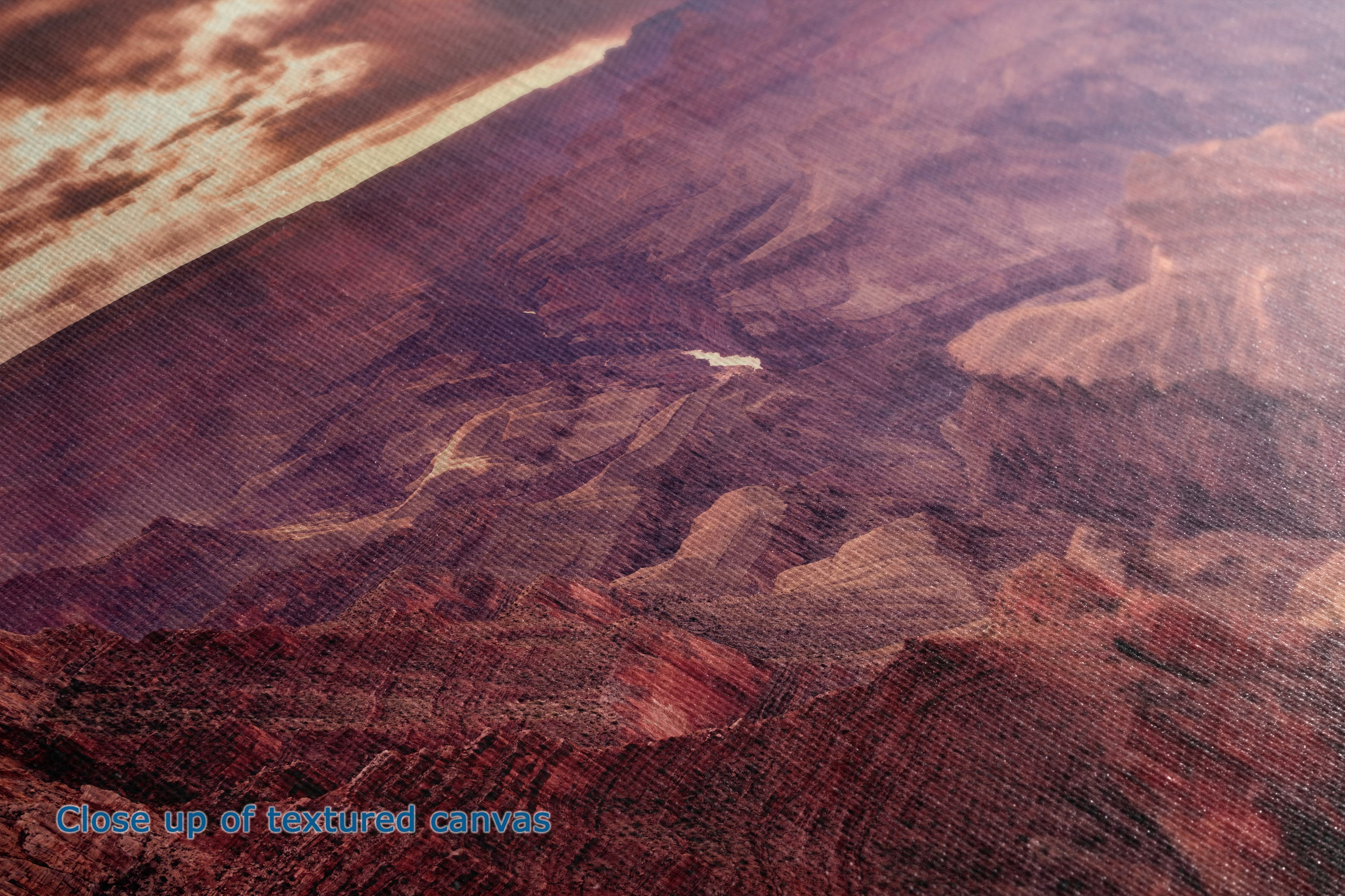 Close-up of a textured canvas print depicting an aerial canyon landscape in warm reddish-brown and purple tones, with a small blue text overlay in the lower-left corner reading 'Close up of textured canvas'.