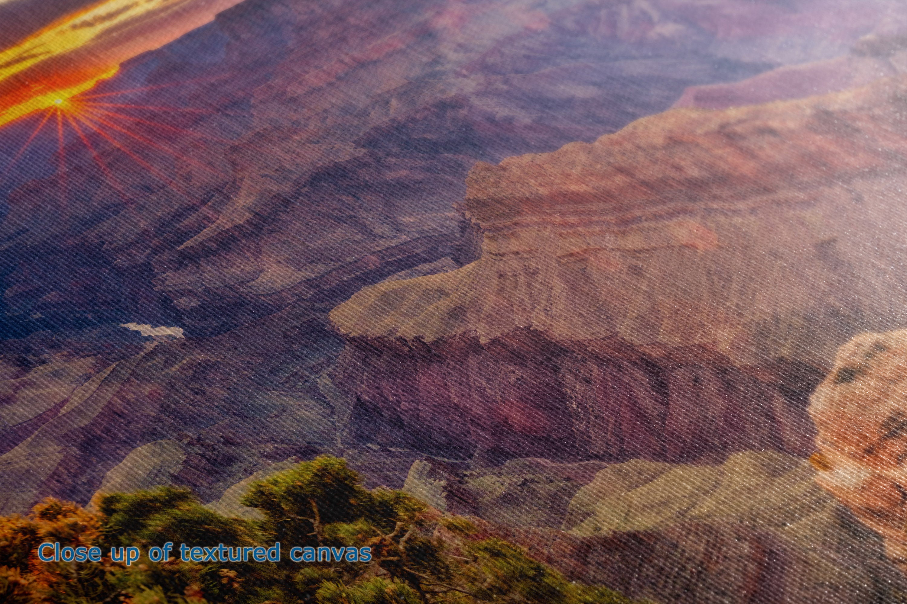 Close-up of a textured canvas print depicting a canyon landscape at sunset, with layered reddish-brown rock formations and a bright sunburst in the upper left.