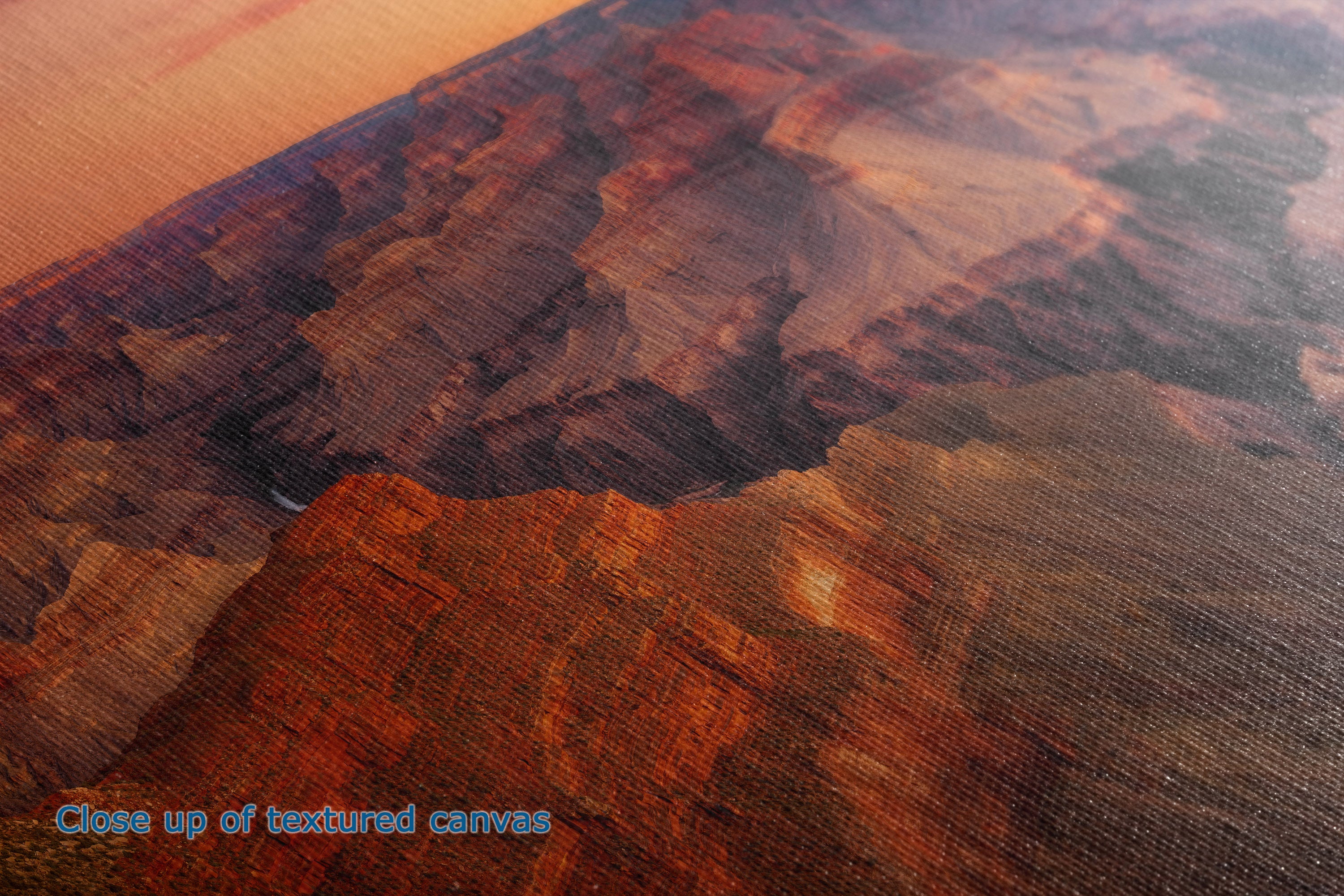 Close-up of a textured canvas print depicting a desert canyon landscape in warm orange, brown, and reddish tones, with blue text in the lower-left corner reading 'Close up of textured canvas'.