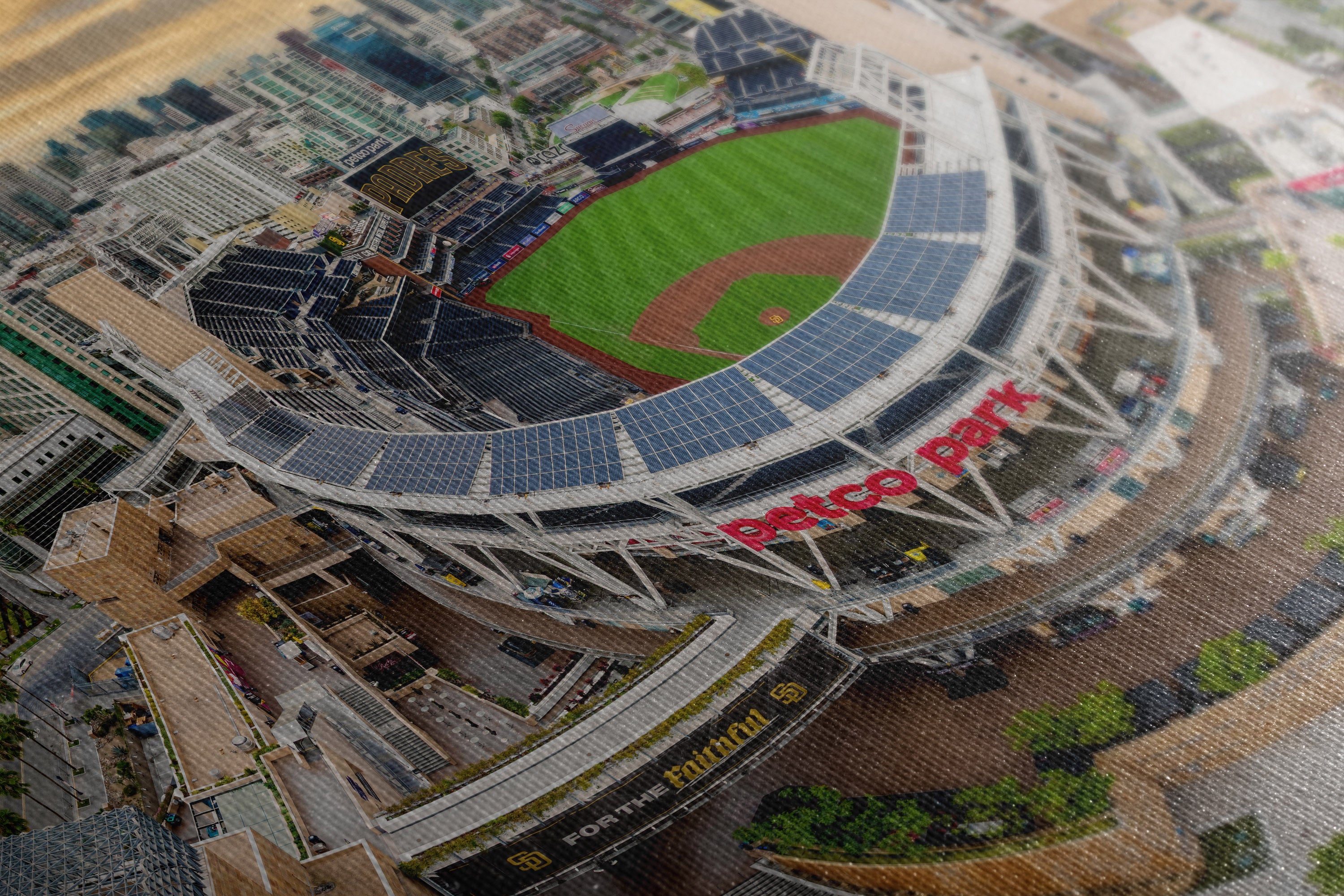 A close-up of a printed or illustrated stadium scene showing a baseball field with a large oval stadium, solar panel-covered upper deck, surrounding city buildings, and red 'Petco Park' lettering on the front.