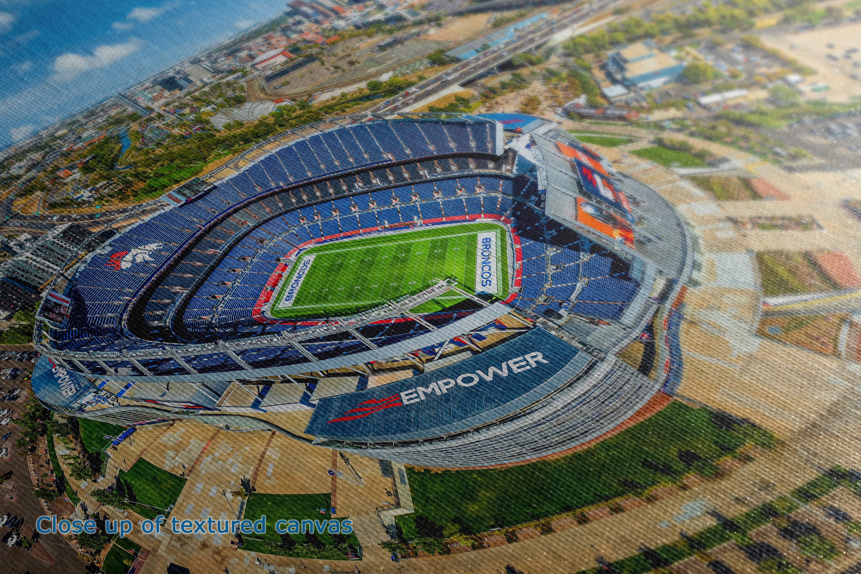 A close-up of the detail and texture of a canvas wall art print depicting an aerial view of a football stadium with blue seating, a green field, and surrounding urban landscape. A small blue text overlay at the bottom left reads 'Close up of textured canvas'.