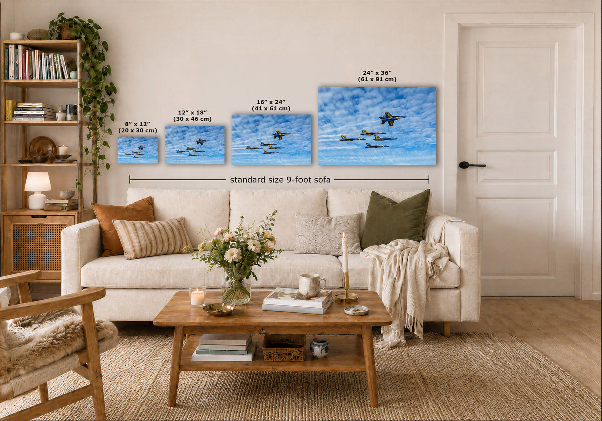 A staged living room showing four blue sky canvas prints above a white sofa, with size labels for each print and a note reading 'standard size 9-foot sofa.' The room includes a wooden coffee table, books, a chair, rug, and a white door.