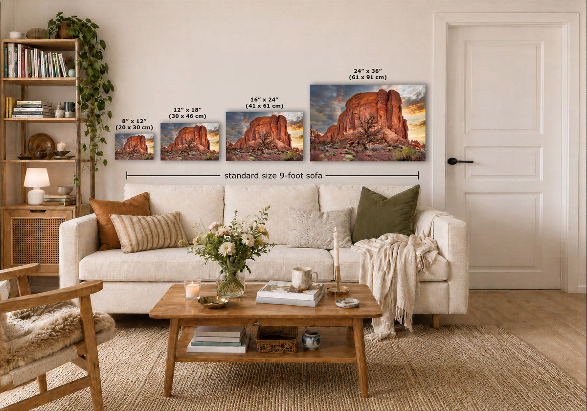 A styled living room showing four landscape art prints above a white sofa, with size labels overlaid for each print and a note reading 'standard size 9-foot sofa.' The scene includes a wooden coffee table, books, a chair, and a door in a real interior environment.