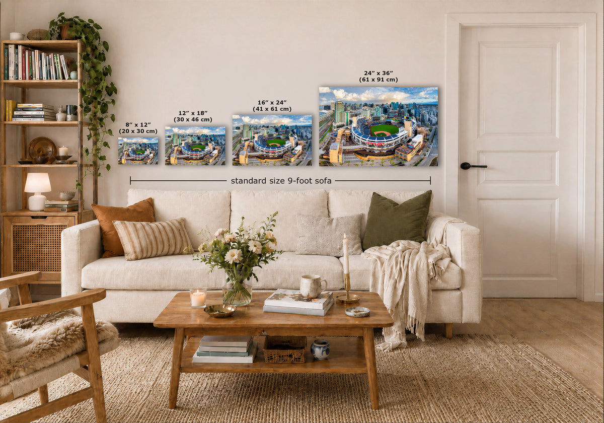 Four wall art prints showing stadium scenes are displayed above a white sofa in a styled living room. White dimension labels and text indicate sizes: 8" x 12" (20 x 30 cm), 12" x 18" (30 x 46 cm), 16" x 24" (41 x 61 cm), and 24" x 36" (61 x 91 cm), with a note reading standard size 9-foot sofa.