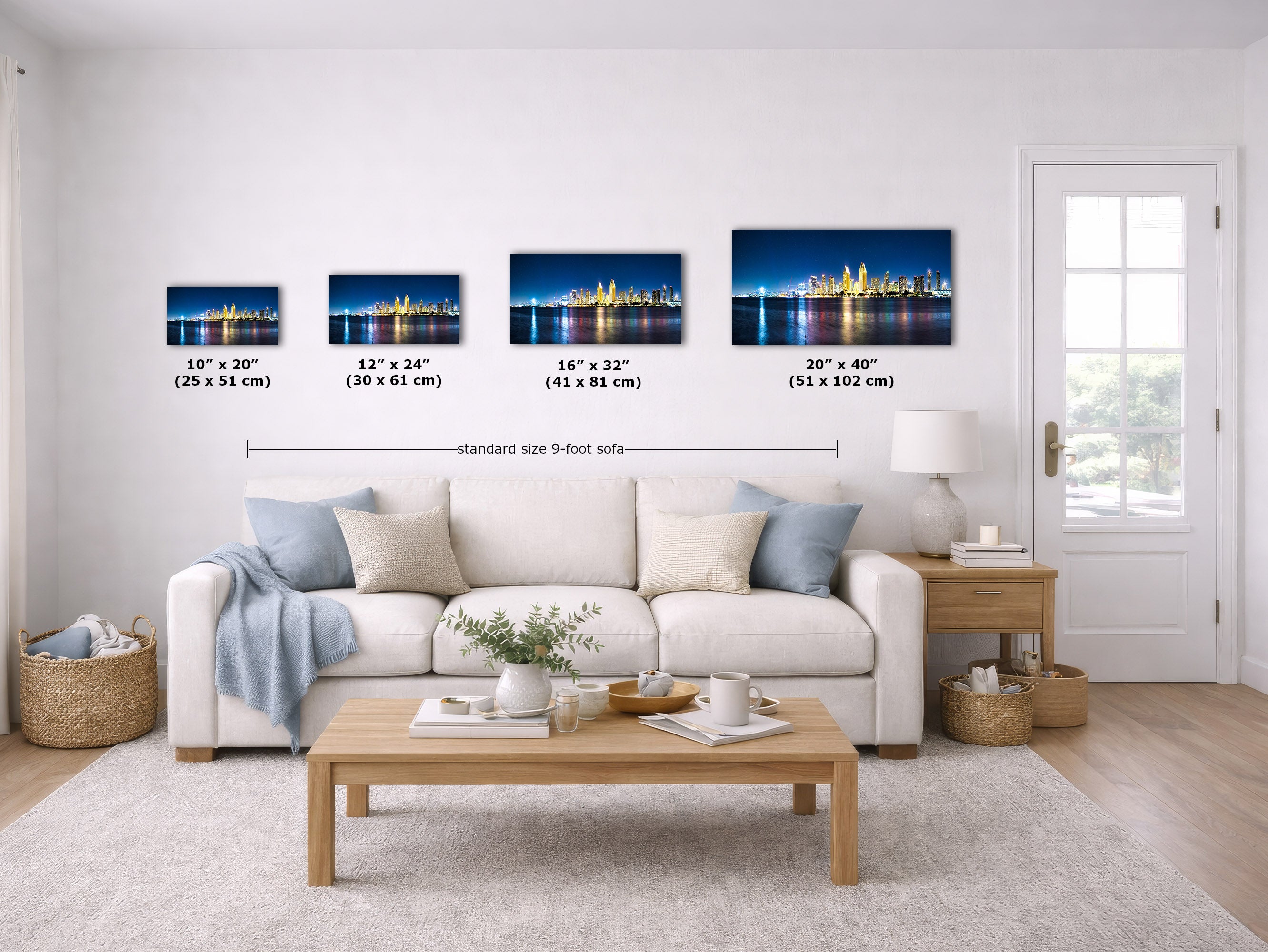 A staged living room showing four framed wall art prints above a white sofa, each with size labels underneath. The prints depict a nighttime city skyline with blue water reflections.