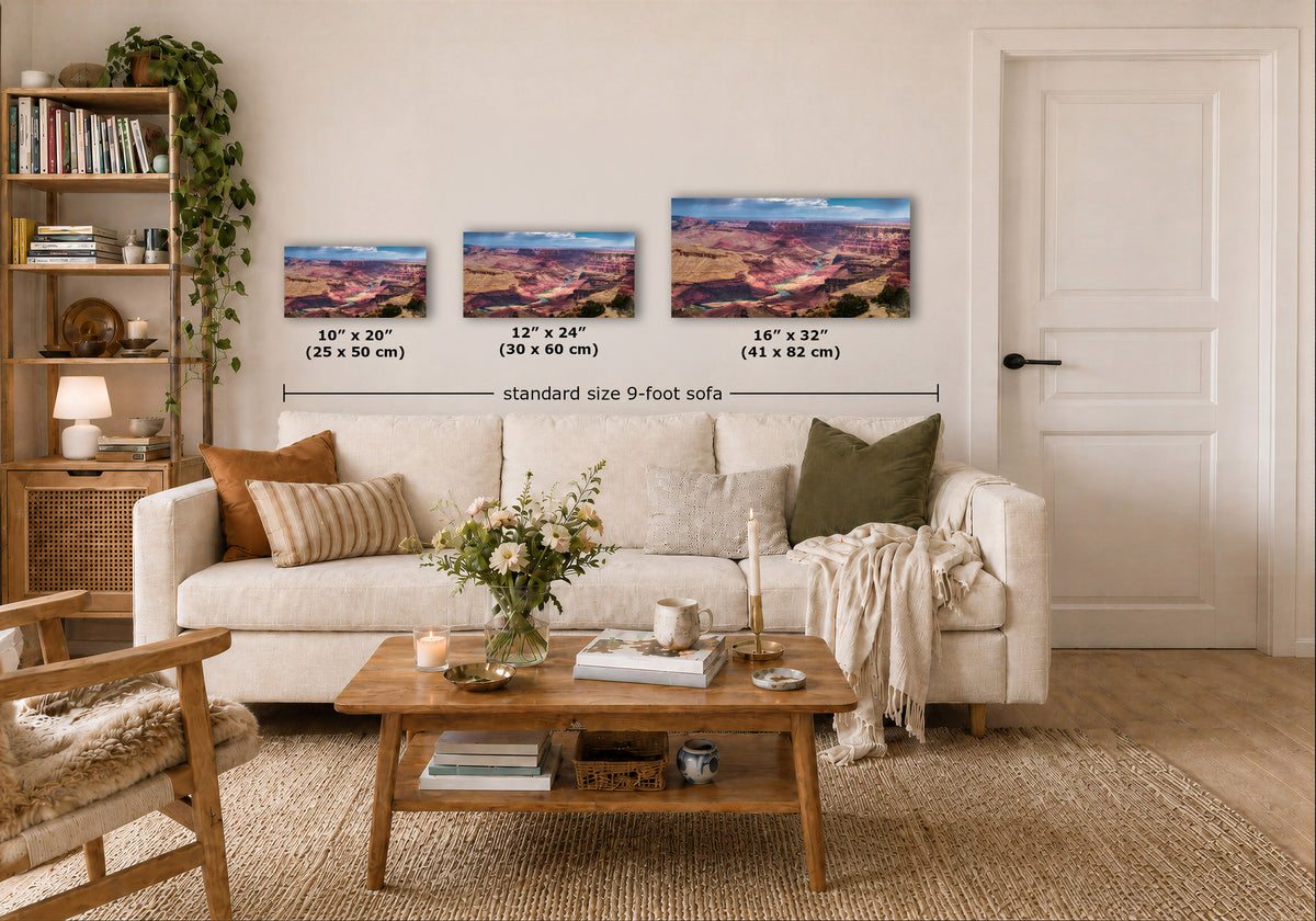 Three wall art prints showing colorful sunset landscapes are displayed above a white sofa in a styled living room. Text labels indicate the dimensions of each print: 10 x 20 inches, 12 x 24 inches, and 16 x 32 inches, with a total width note reading standard size 9-foot sofa.