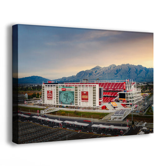 a large stadium with a red roof and a large screen displaying a logo, set against a backdrop of mountains and a cloudy sky.