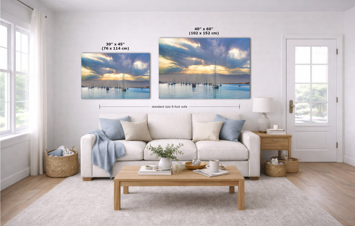 A staged living room with a white sofa, coffee table, and two wall art prints above the couch. The prints show a sunrise over calm harbor with many boats, and overlaid text indicates two sizes: 30 x 45 inches and 40 x 60 inches.