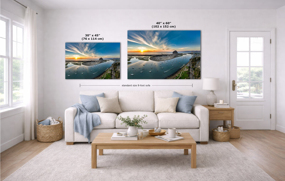 A staged living room with a white sofa, coffee table, and two landscape wall art prints above the couch. The prints show a coastal sunset scene with blue water and warm sky tones, with size labels and dimension lines overlaid.