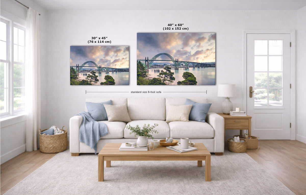 A staged living room with a white sofa, coffee table, and two landscape wall art prints above the couch. The prints show a colorful sunset sky over water with a bridge, and size labels are overlaid on each print.