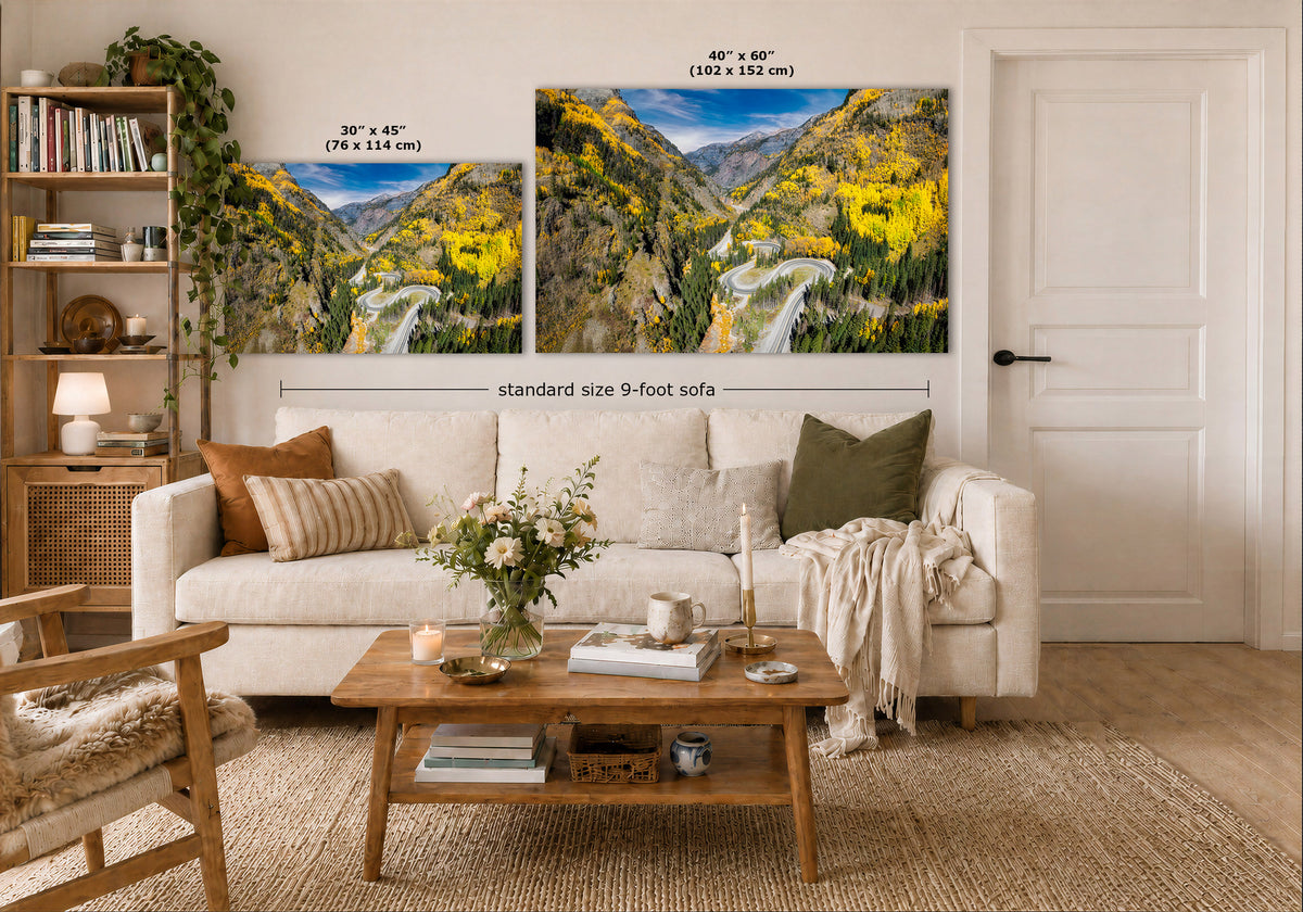 A styled living room showing two landscape art prints above a white sofa. The prints depict a mountain valley with yellow wildflowers under a blue sky.