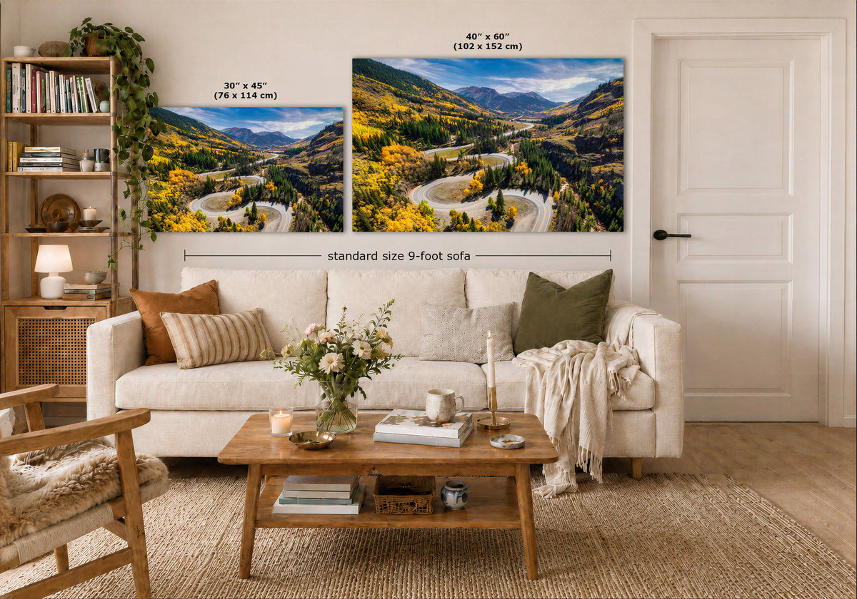 Two large landscape wall art prints displayed above a white sofa in a styled living room. The prints show mountain valleys with winding roads and autumn foliage.