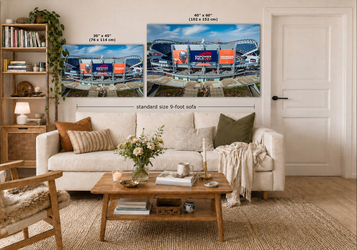 A staged living room shows a Broncos Empower Field Wall Art set featuring two stadium scenes mounted above a white sofa. The image includes size callouts for the prints: one 30 x 45 in (76 x 114 cm) and one 40 x 60 in (102 x 152 cm), with a note reading standard size 9-foot sofa.