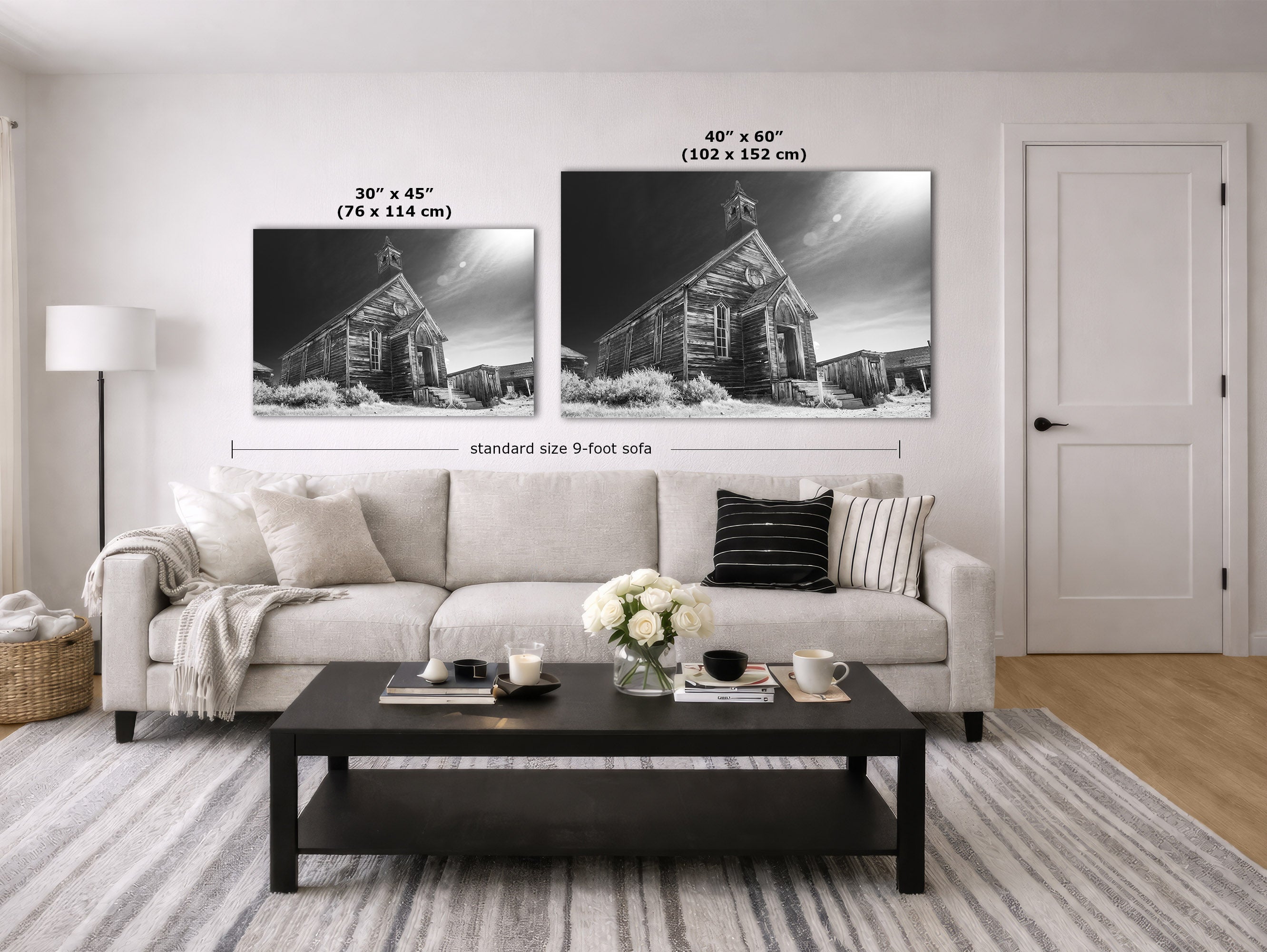 A staged living room showing two black-and-white wall art prints above a light sofa. The prints depict a rustic wooden church building in a snowy landscape.