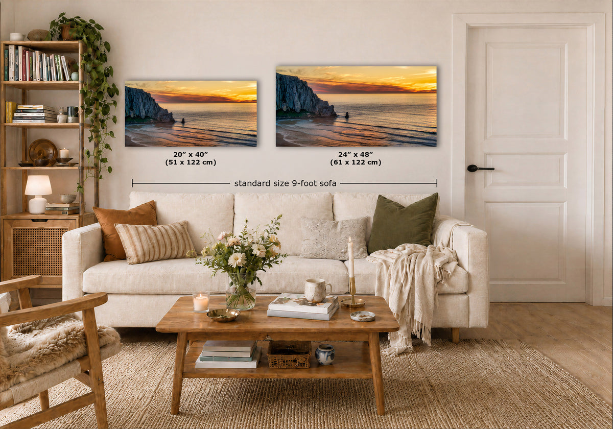 Two wall art prints showing a coastal sunset scene are displayed above a white sofa in a styled living room. Black dimension lines and text indicate the sizes of each print: 20 x 40 inches (51 x 122 cm) and 24 x 48 inches (61 x 122 cm), with a note for a standard size 9-foot sofa.