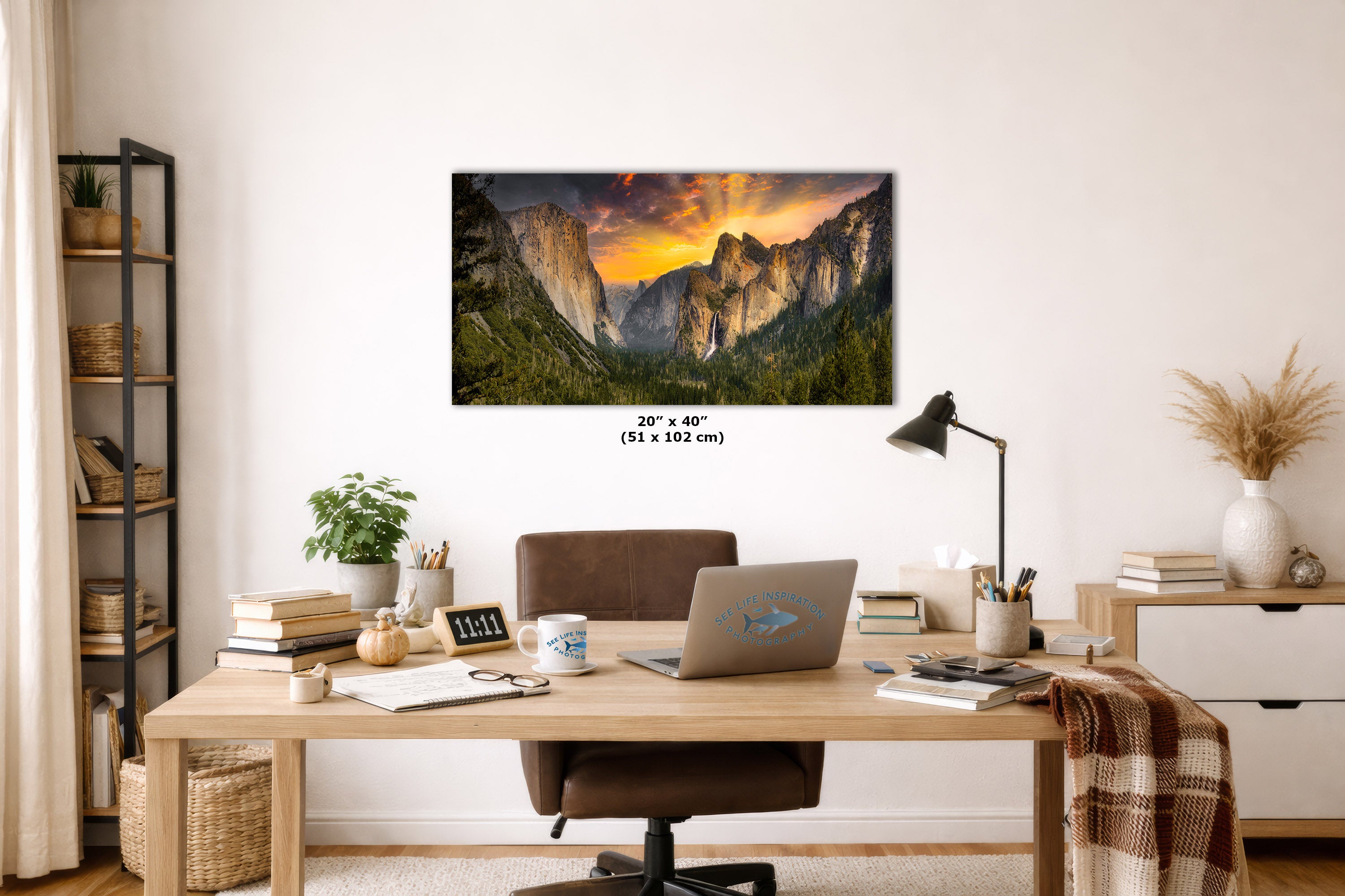 A framed wall art print showing a mountain landscape at sunset hangs above a wooden desk in a styled home office. The print is the product being sold, but the image includes a real interior environment with desk, chair, lamp, books, and decor.