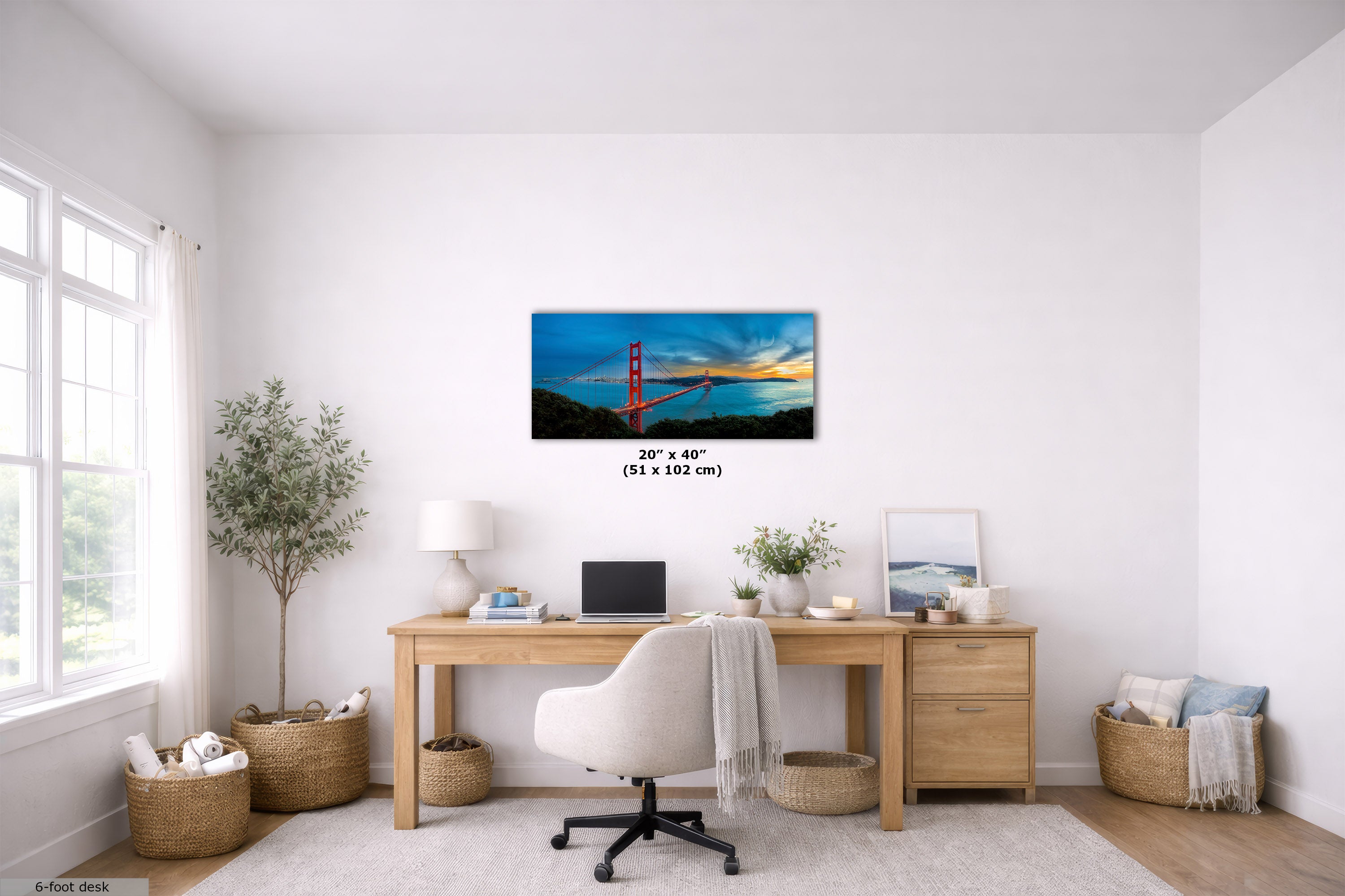 A framed wall art print of the Golden Gate Bridge over water at sunset, shown hanging above a wooden desk in a bright, modern home office. The print has a black frame and size text reading 20 x 40 (51 x 102 cm) below it.