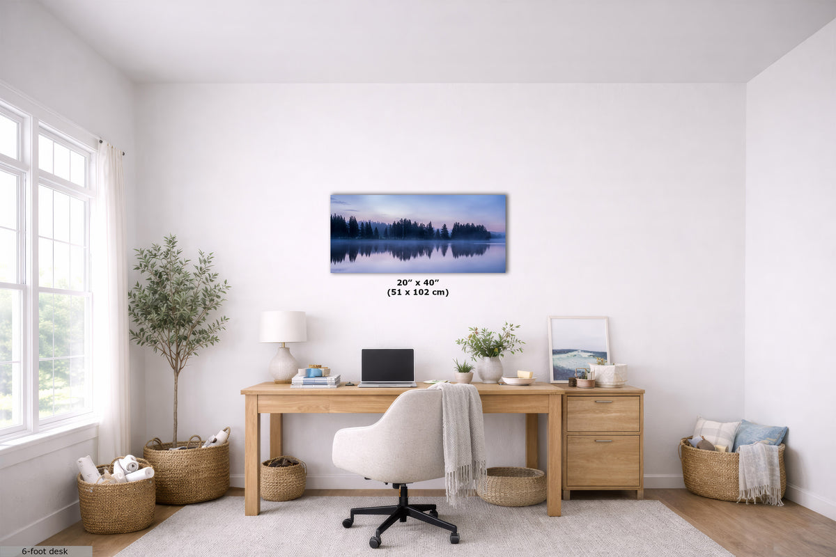 A framed horizontal landscape art print depicting a misty blue forest reflected in calm water, displayed on a white wall above a wooden desk in a bright, modern home office. The print includes a size overlay indicating 20" x 40" (51 x 102 cm).