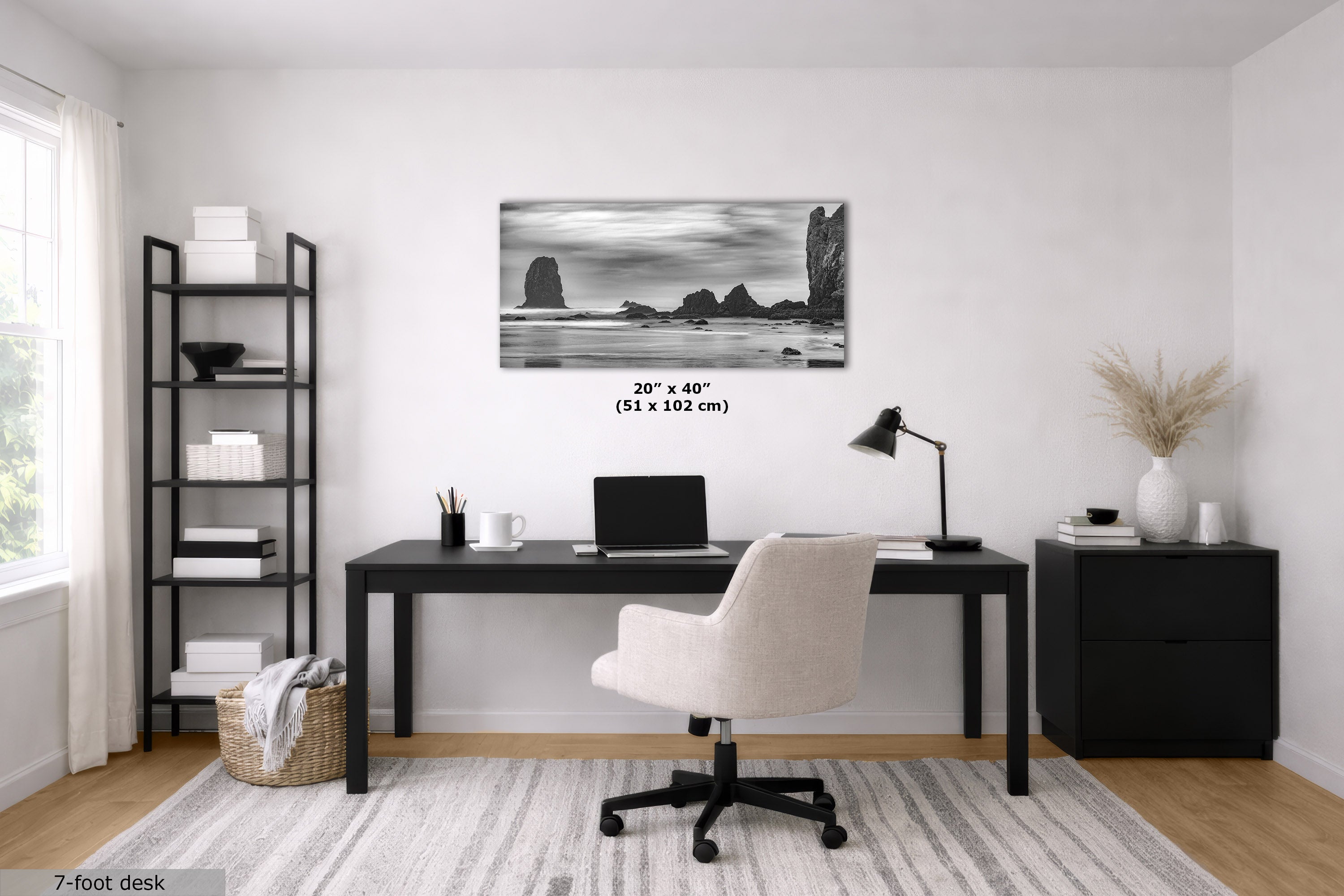 A black-framed horizontal wall art print depicting a black and white coastal rock formation, displayed in a modern home office setting above a black desk and white swivel chair.