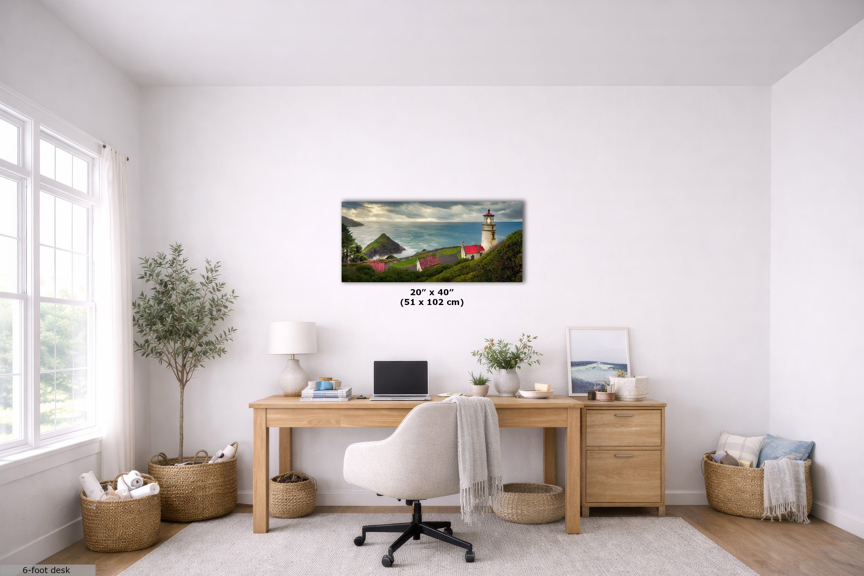 A framed wall art print depicting a coastal lighthouse scene with green hills, red-roofed buildings, and blue water, displayed above a wooden desk in a bright, modern home office. The print includes size text reading 20 x 40 in (51 x 102 cm) and a small scale graphic.