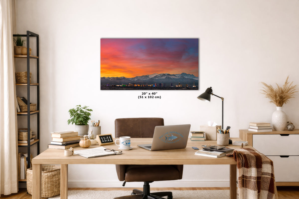 A rectangular wall art print showing a colorful sunset over a mountain range, displayed above a wooden desk in a styled home office setting. The print includes size text reading 20" x 40" (51 x 102 cm) below it.