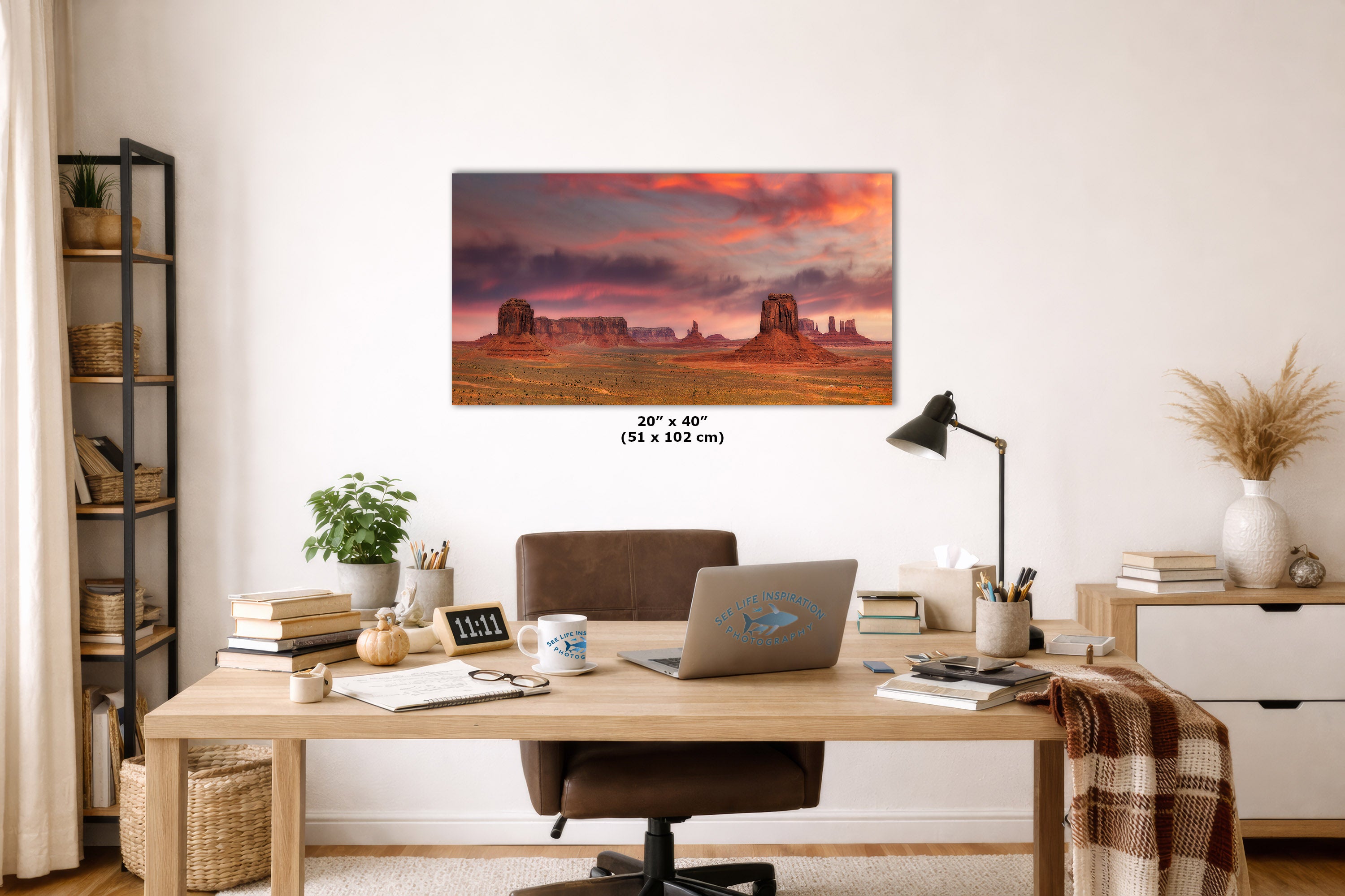 A wall art print depicting a desert landscape at sunset hangs above a wooden desk in a styled home office. The print shows orange and pink sky tones with dark rock formations.