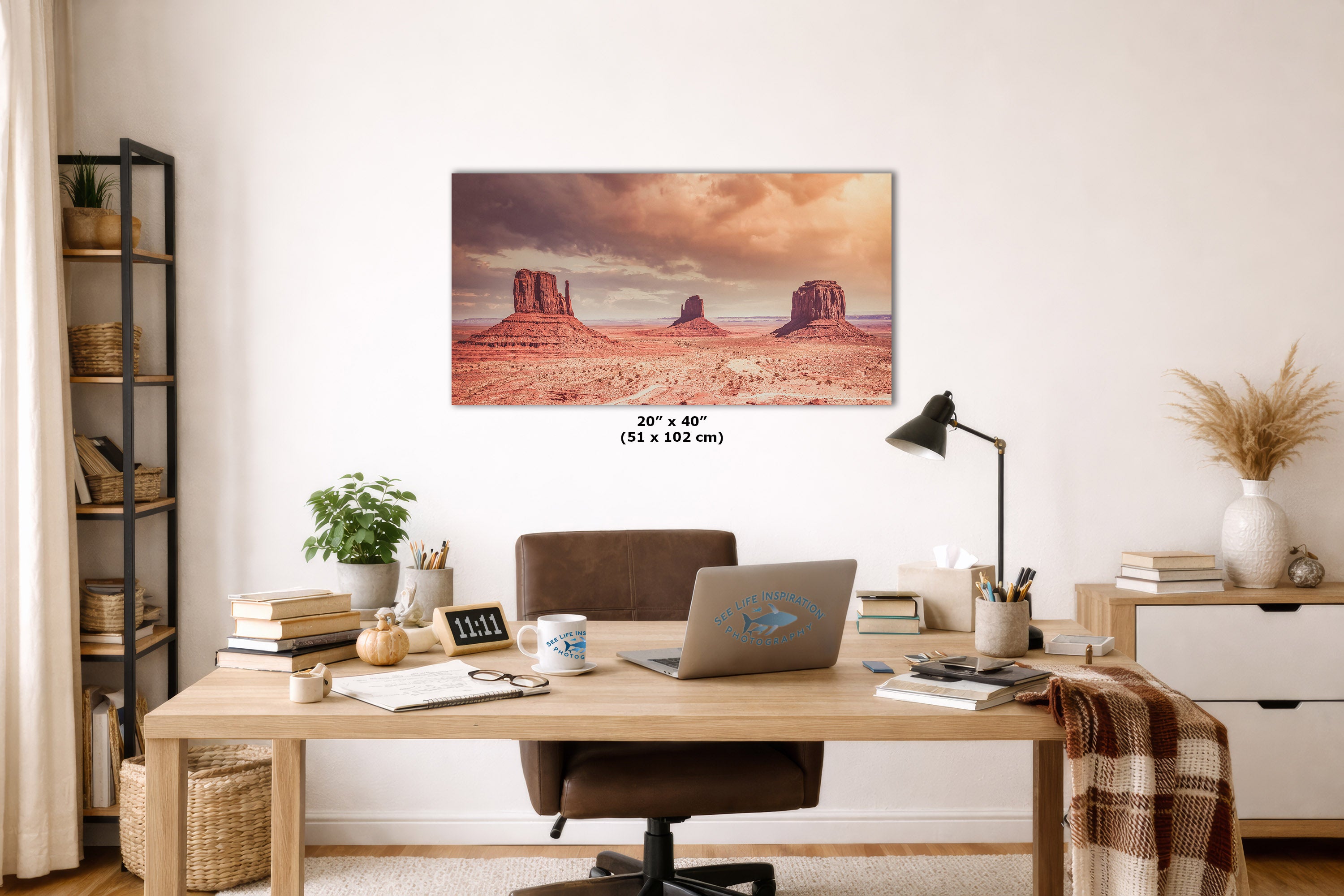 A framed wall art print depicting a desert landscape with red rock buttes under a dramatic sky, displayed above a wooden desk in a styled home office. The print includes size text reading 20 x 40 (51 x 102 cm) below it.
