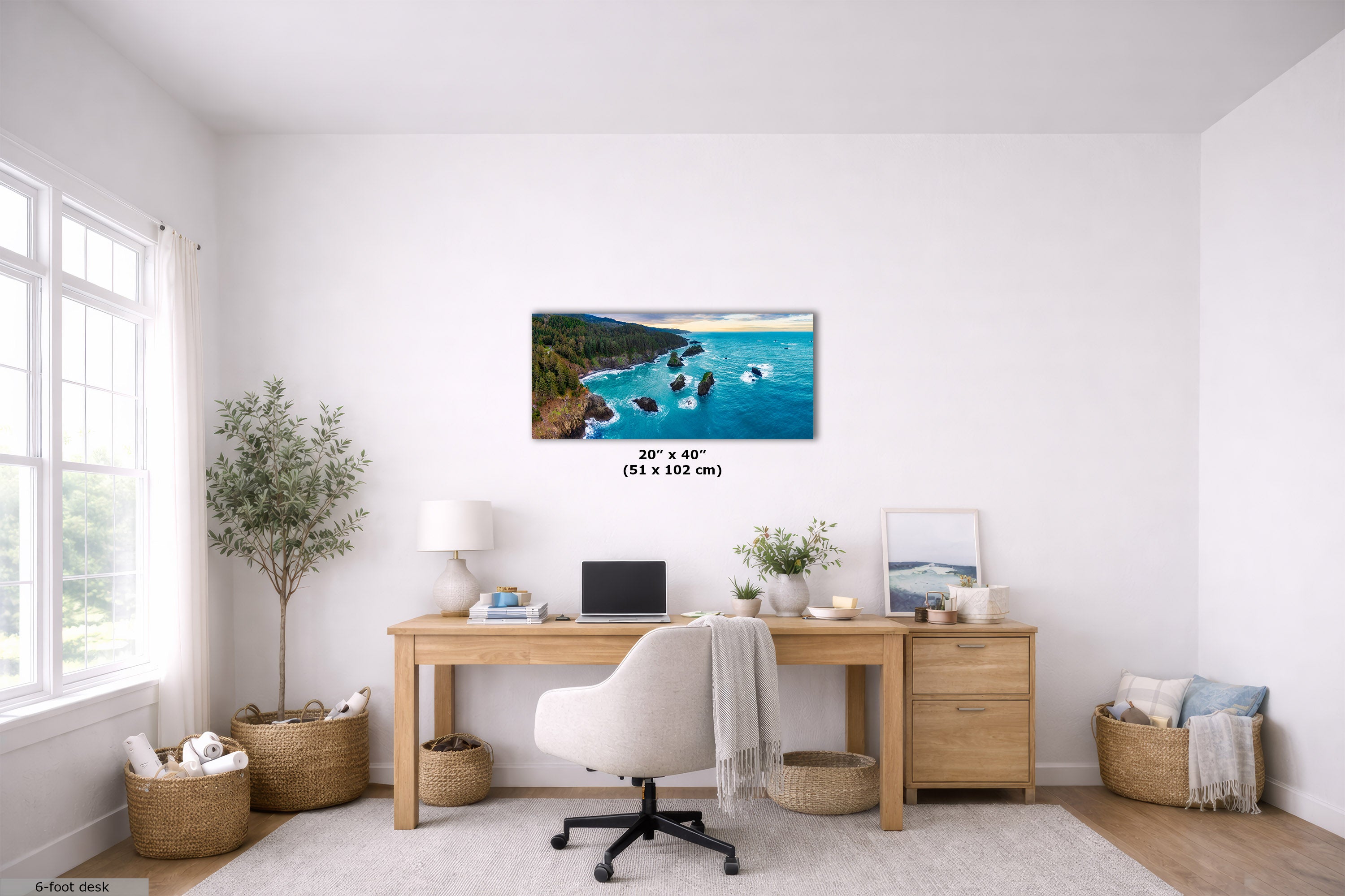 A framed coastal landscape wall art print hangs above a wooden desk in a bright, modern home office. The print shows turquoise water, rocky shoreline, and green cliffs.