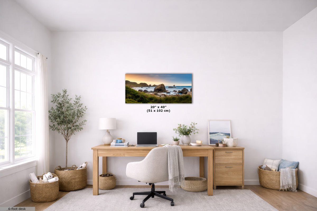 A coastal landscape wall art print hangs above a wooden desk in a bright, modern home office. The print shows a rocky shoreline at sunset with blue water and green cliffs.