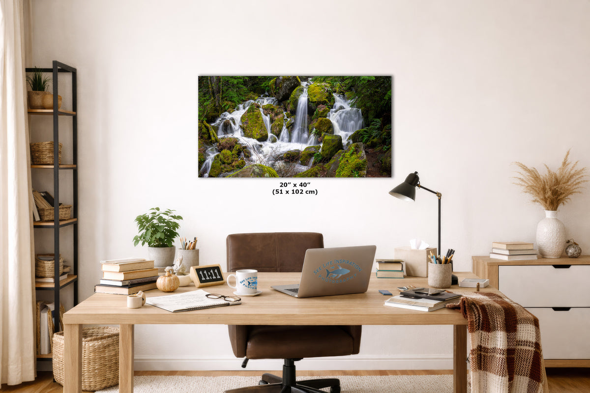 A framed wall art print showing a lush waterfall scene in a real home office setting, mounted above a wooden desk with a laptop, lamp, books, and chair. The print includes size text reading 20" x 40" (51 x 102 cm).