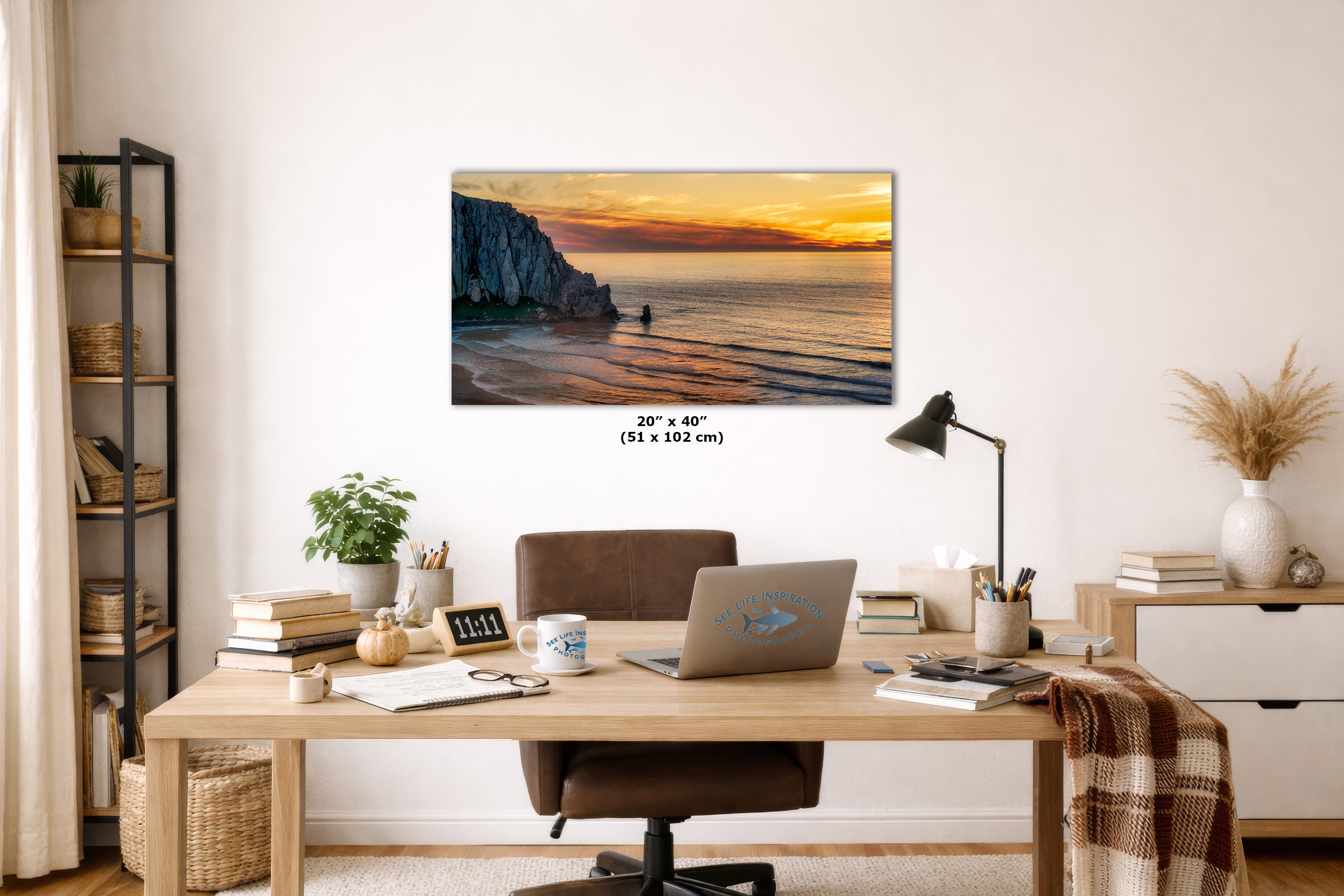A wall art print showing a coastal sunset scene with cliffs and ocean, displayed above a wooden desk in a styled home office. The print includes size text reading 20" x 40" (50 x 102 cm).