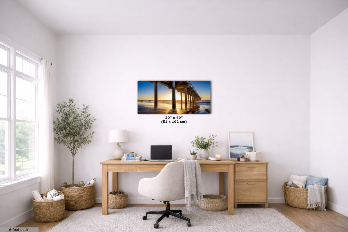 A wall art print depicting a pier over water at sunset, displayed in a bright, modern home office setting above a wooden desk with a white chair.