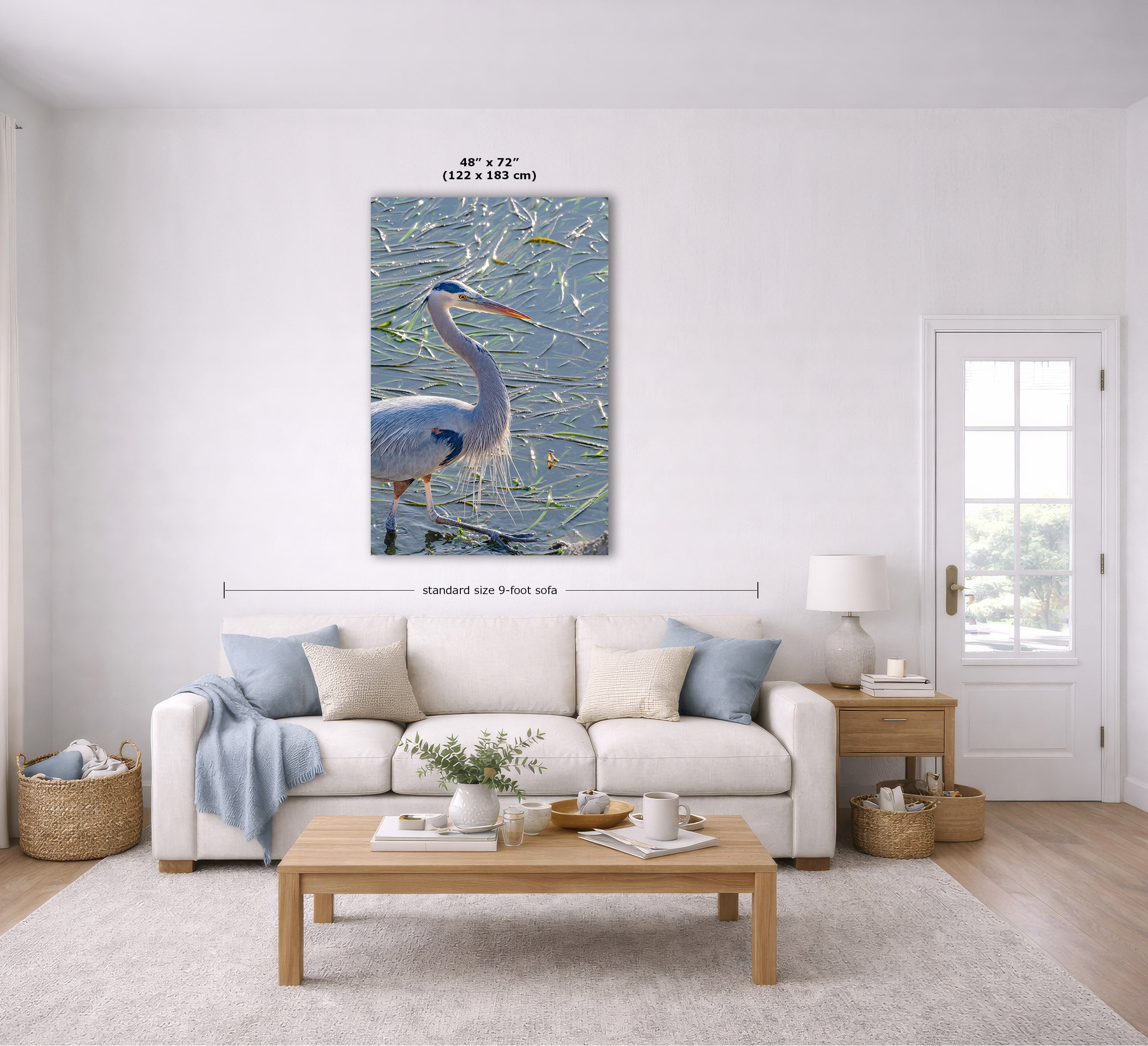 A large vertical canvas print of a blue heron standing in reeds, displayed on a white wall above a beige sofa in a bright, modern living room. The image includes dimension lines indicating the canvas is 48 x 72 inches.