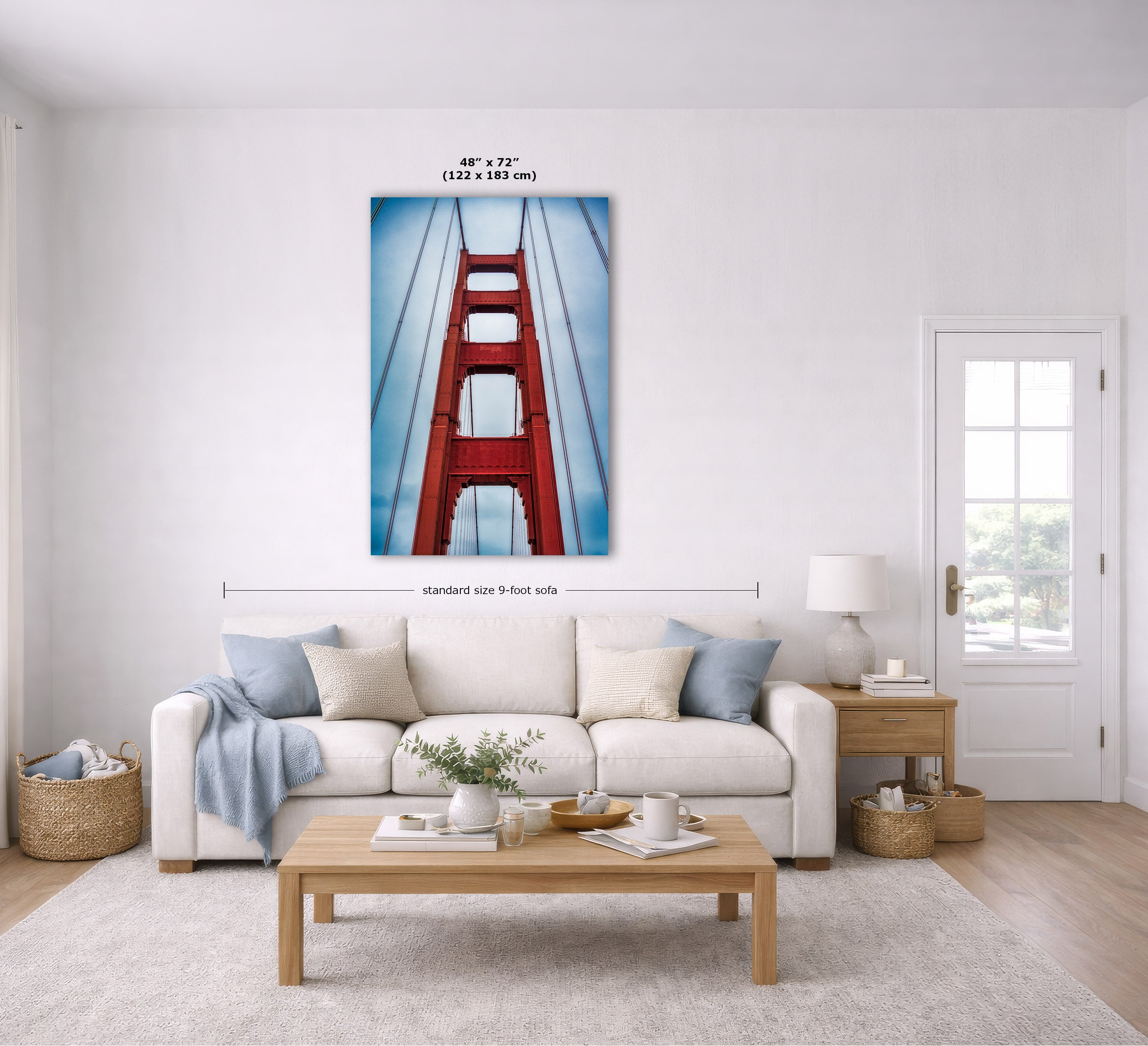 A framed vertical wall art print of the Golden Gate Bridge in red and blue tones, displayed above a white sofa in a bright, modern living room. Measurement lines and text indicate the print is 48 x 72 inches.
