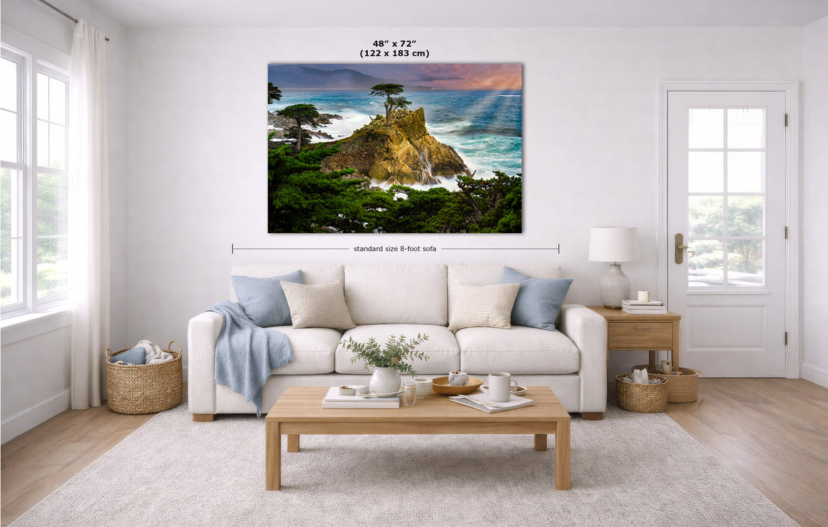 A large coastal landscape wall art print hangs above a white sofa in a bright living room. The artwork shows a rocky cliff with ocean water, green trees, and a colorful sunset sky.